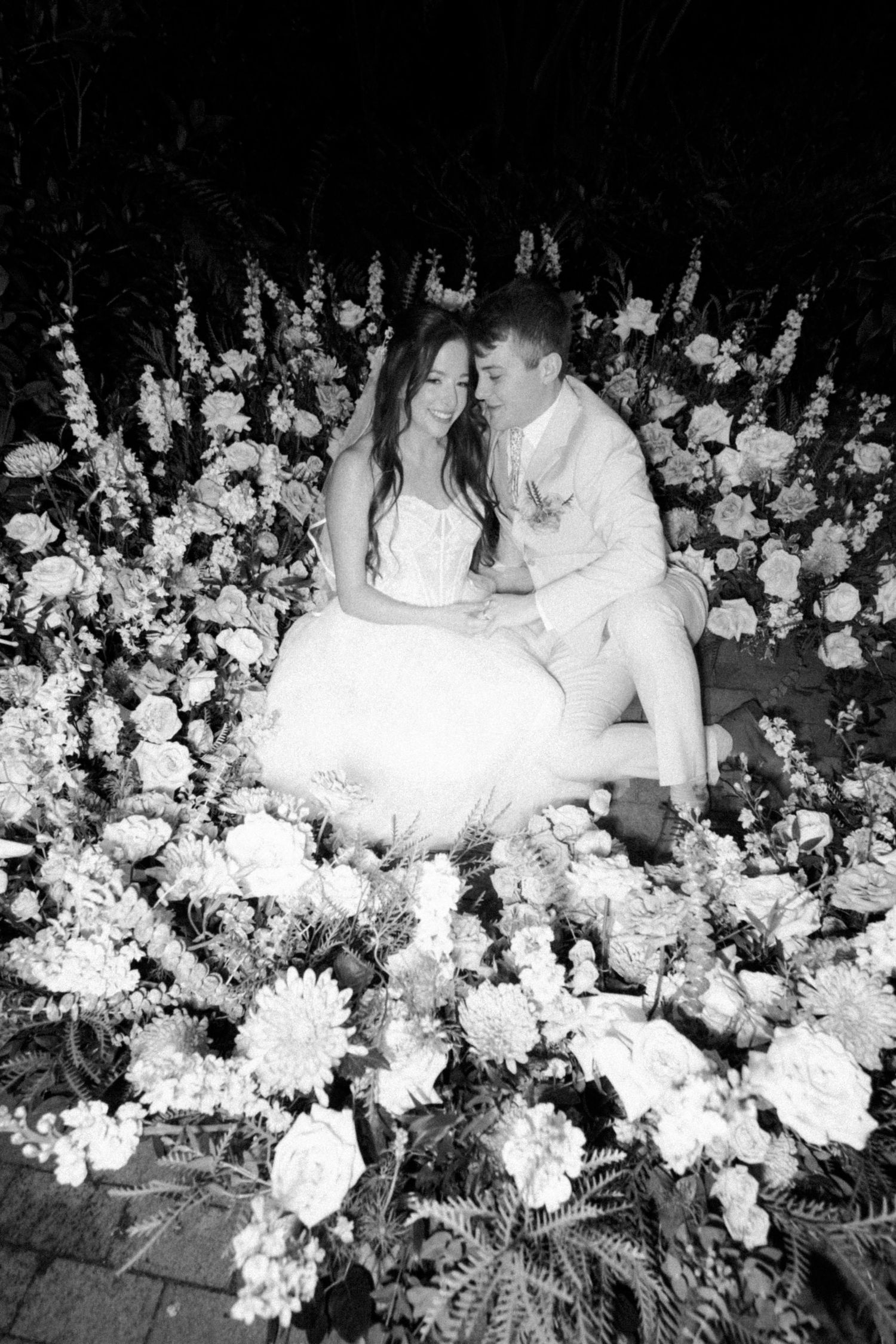 Romantic black and white photo of a couple surrounded by abundant floral arrangements.