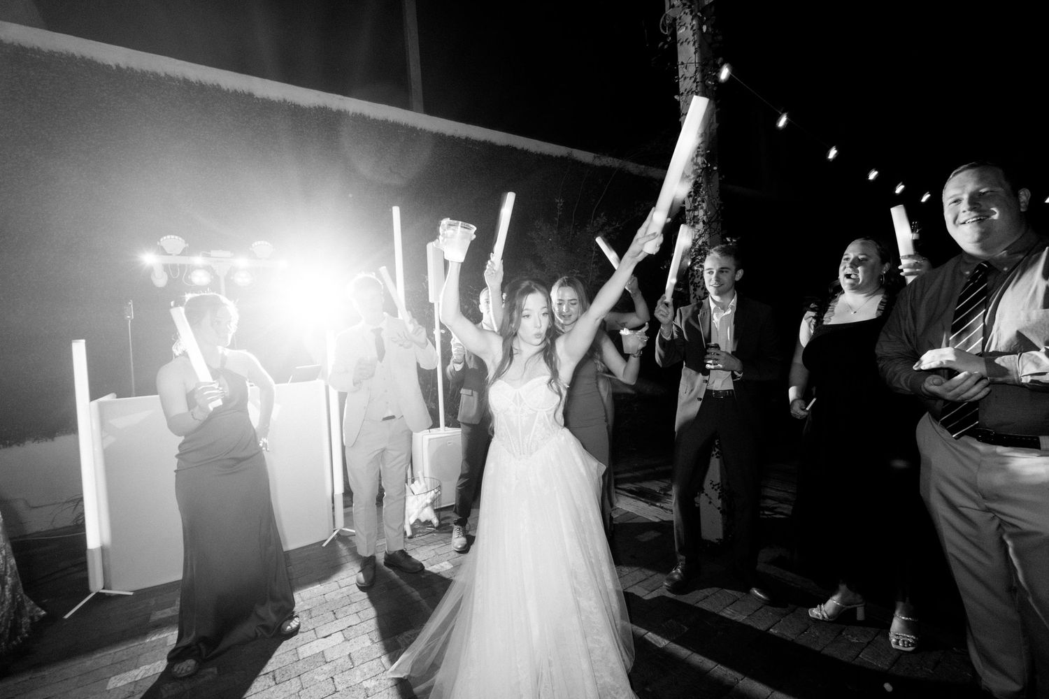 Wedding guests celebrate on the dance floor with raised hands and bright lights in the background.