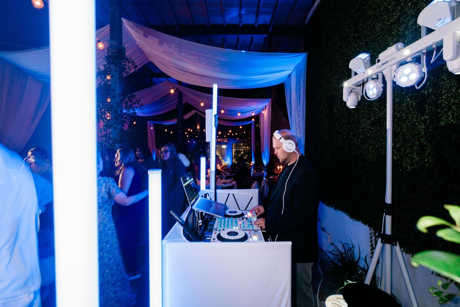 A DJ performs at a nighttime event under white fabric draping and blue lighting at an outdoor venue setup.
