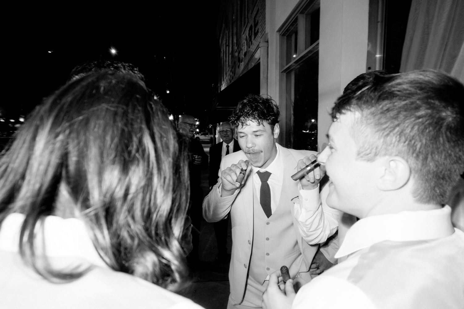 A black and white candid party photograph shows figures engaged in conversation at a nighttime event.