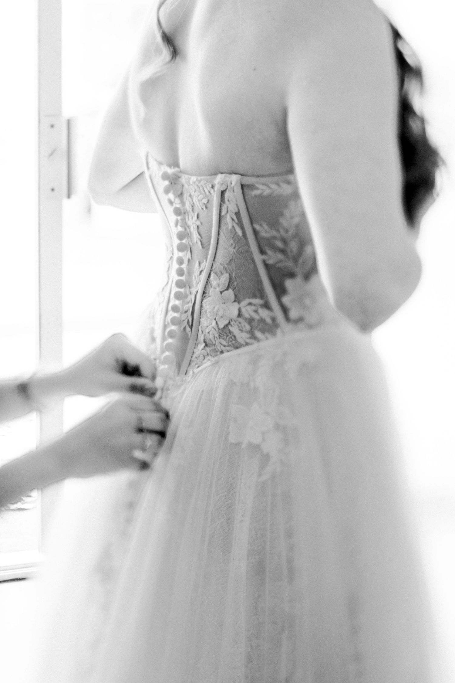 Black and white close-up of a bride getting laced into an ornate corset-style wedding dress bodice.