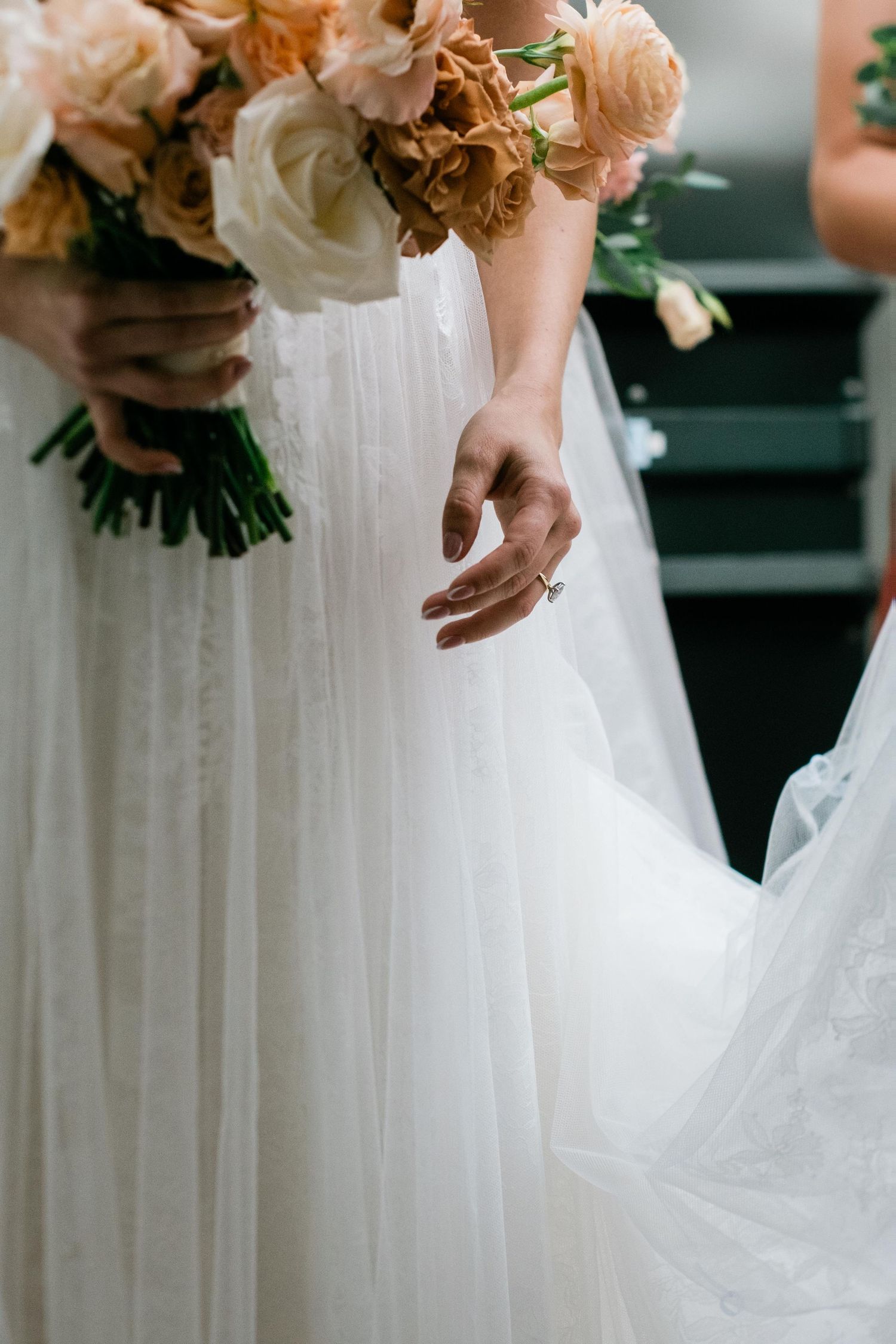 A closeup of a white wedding dress and peach-colored bouquet being held at a wedding ceremony.