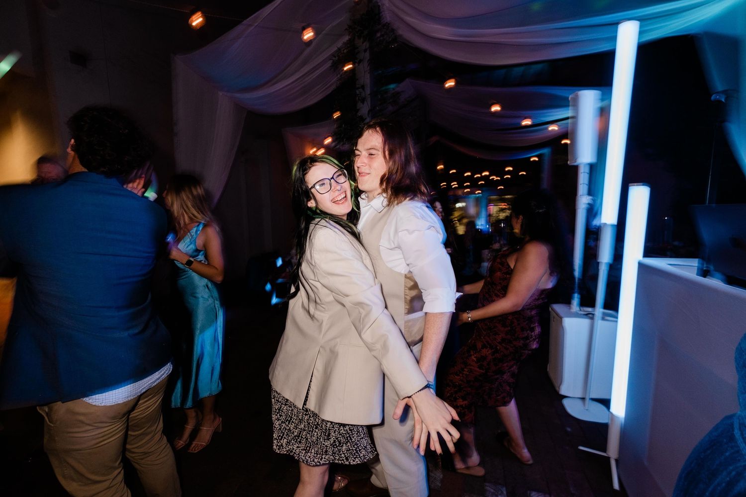 People dancing together in a dimly lit venue with blue mood lighting and decorative ceiling draping.