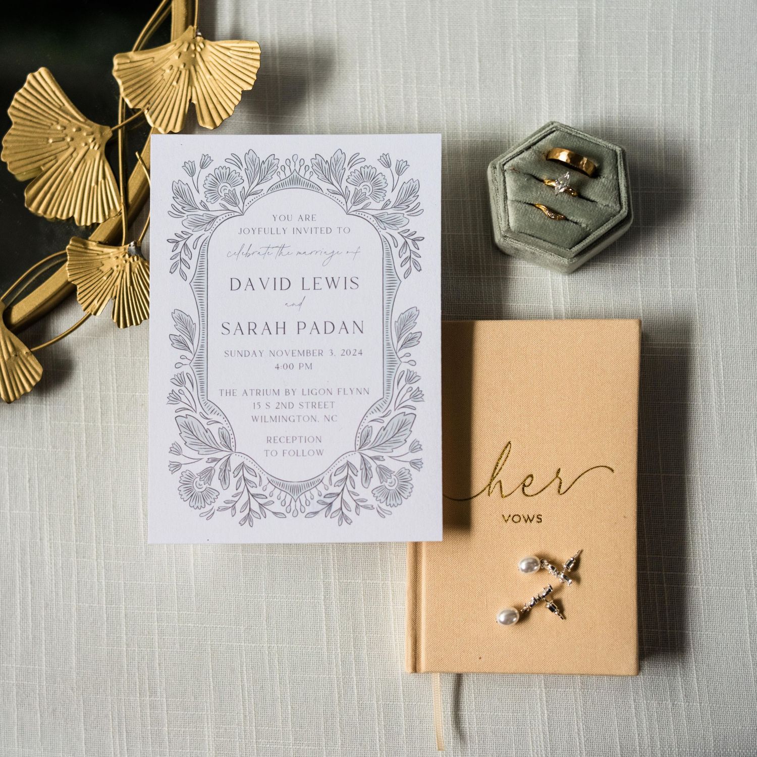 Wedding invitation with gold ginkgo leaf decor, vow notebook, and hexagonal ring box on light gray background.