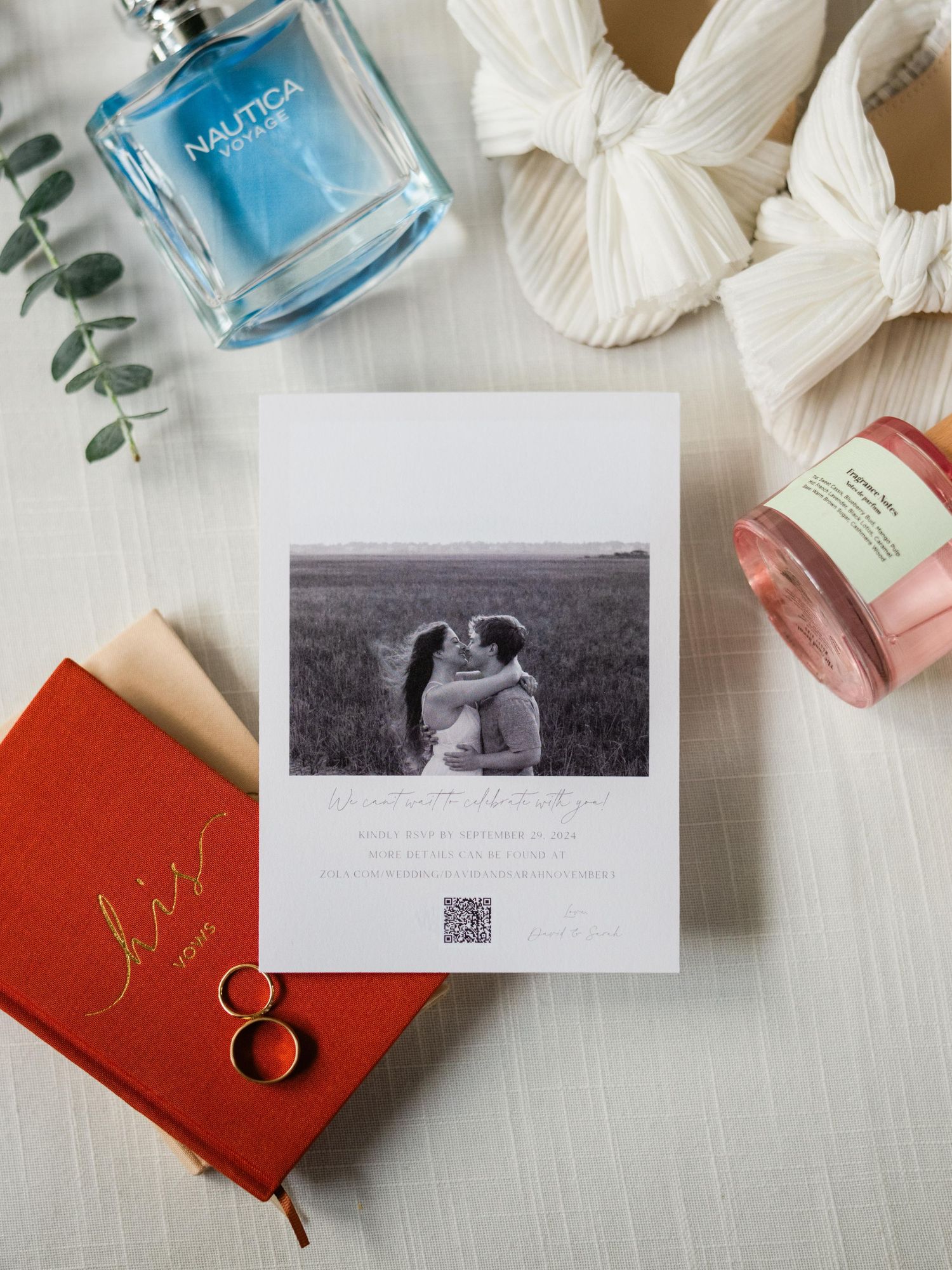 Flatlay of perfume bottles, wedding rings, a red journal, and a black and white photograph on white fabric with eucalyptus.