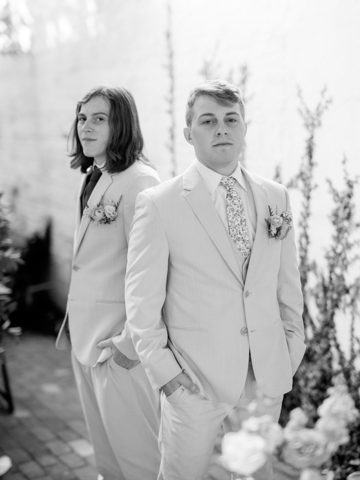 Two figures in matching white suits with boutonnieres pose back to back in a black and white formal portrait.