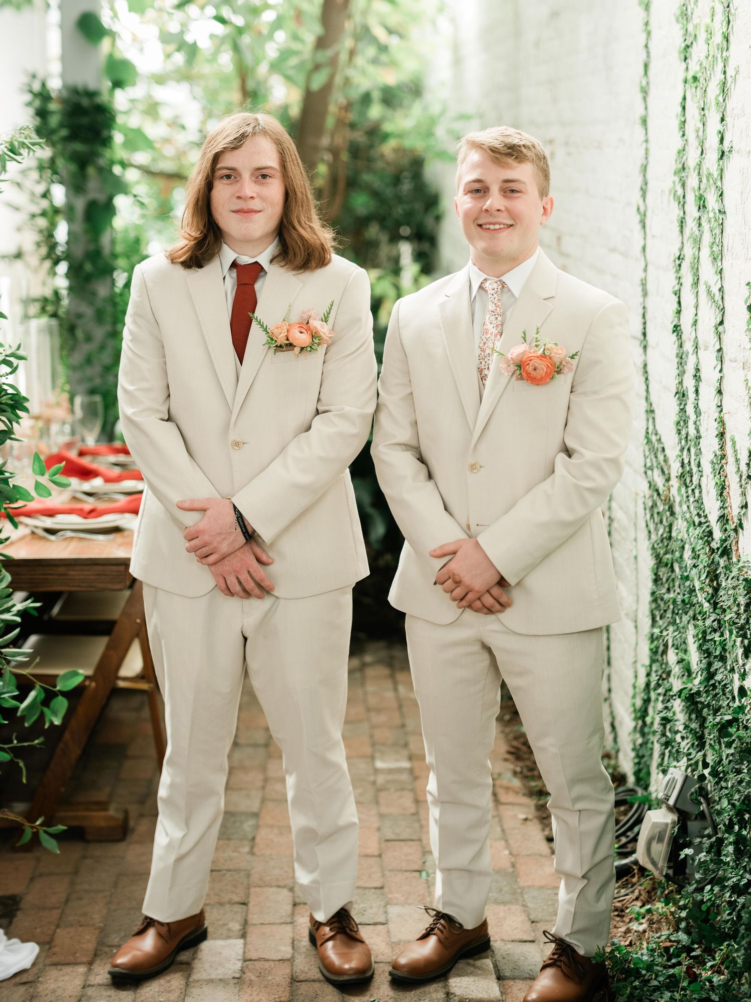 Two people wearing matching cream-colored suits with orange boutonnieres stand together at an outdoor wedding venue.
