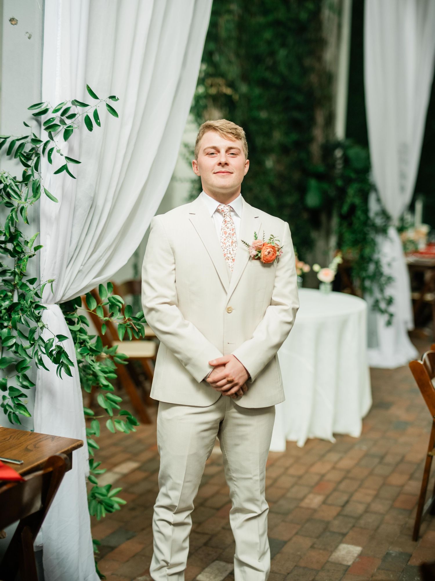 A groom in a light cream suit with a coral boutonniere stands in front of white curtains at an outdoor wedding venue.