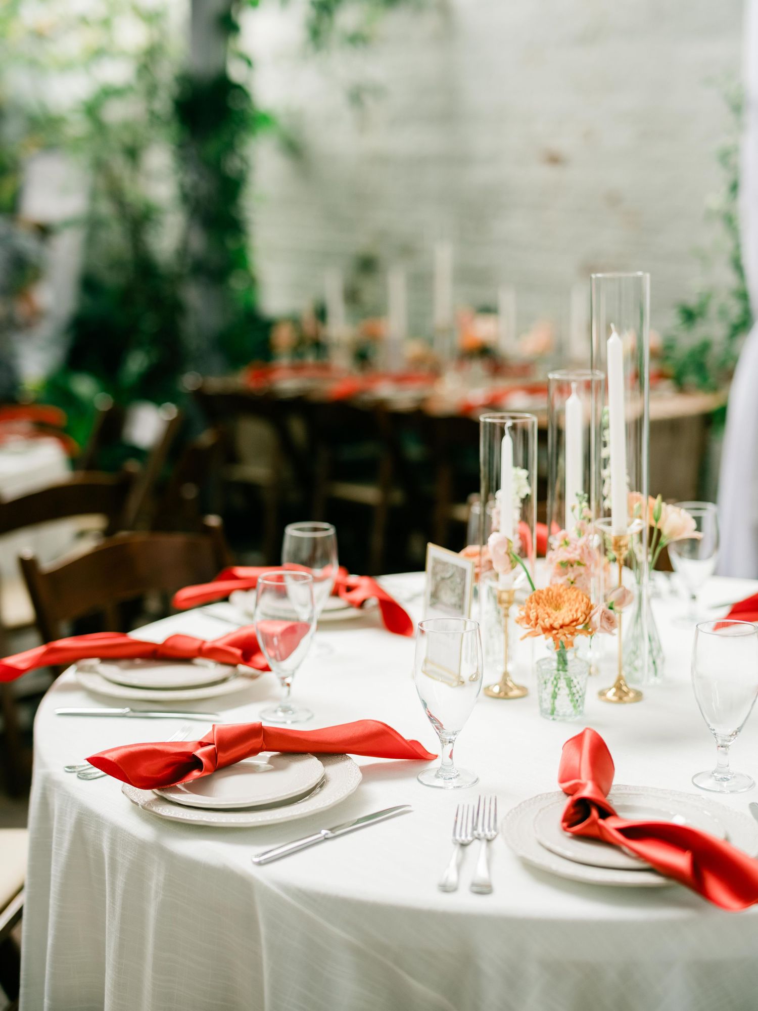 An elegant wedding table setting with red napkins, white linens, and glass candleholders in a garden-style venue.