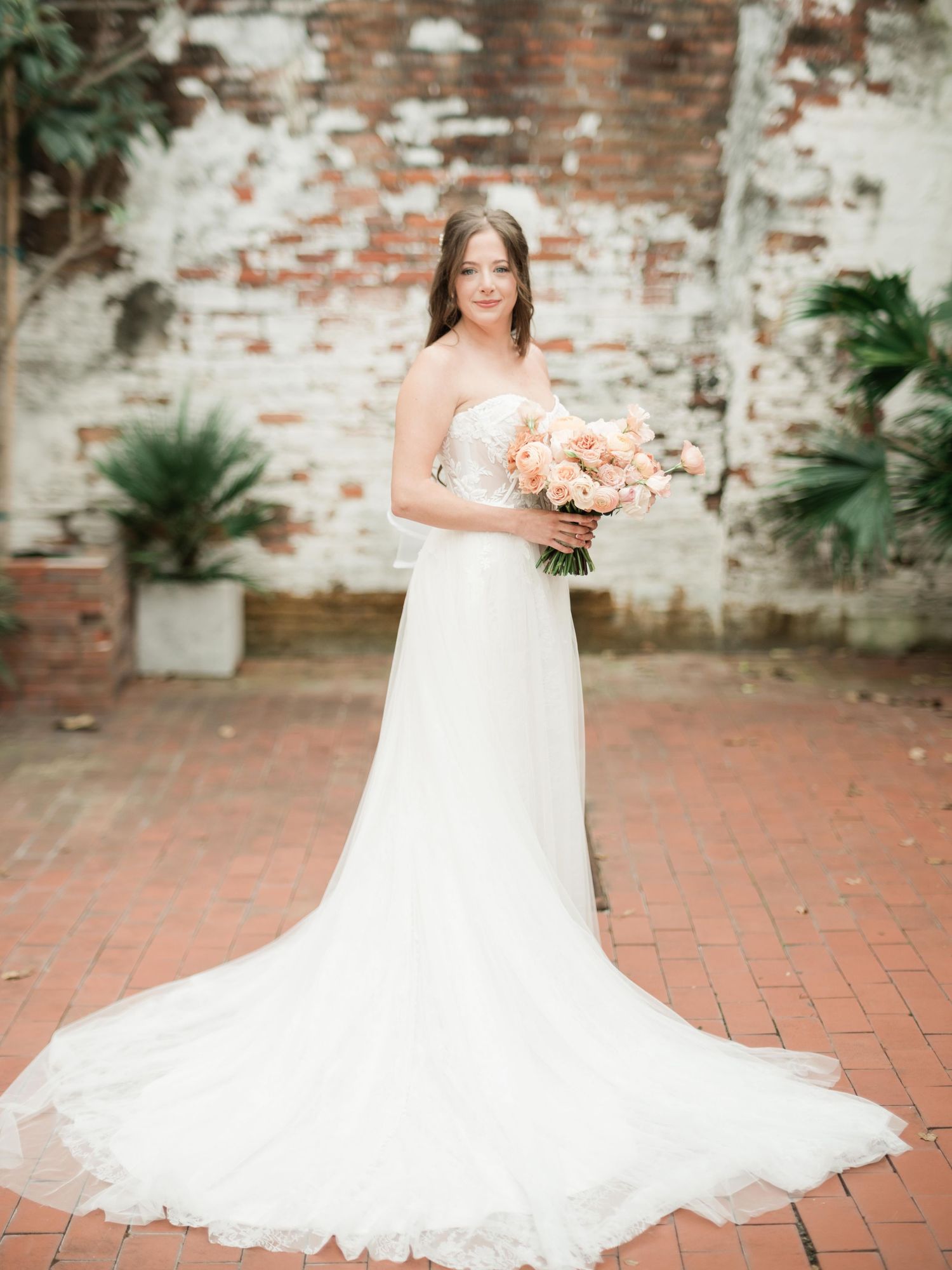 Elegant bridal portrait featuring a flowing white gown and peach bouquet against a rustic brick wall.