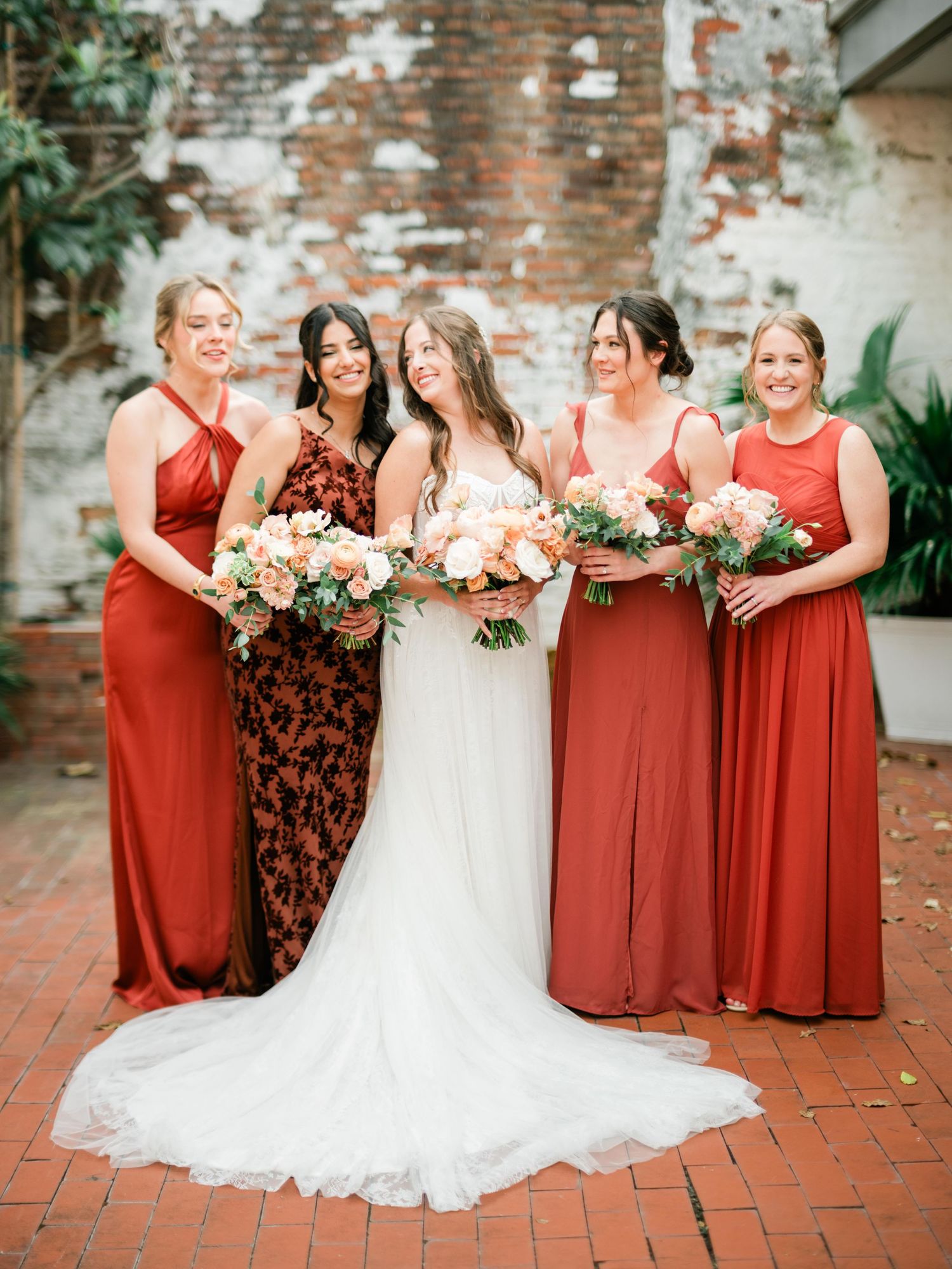 Bridal party in coral and floral dresses poses against weathered brick wall with tropical plants.