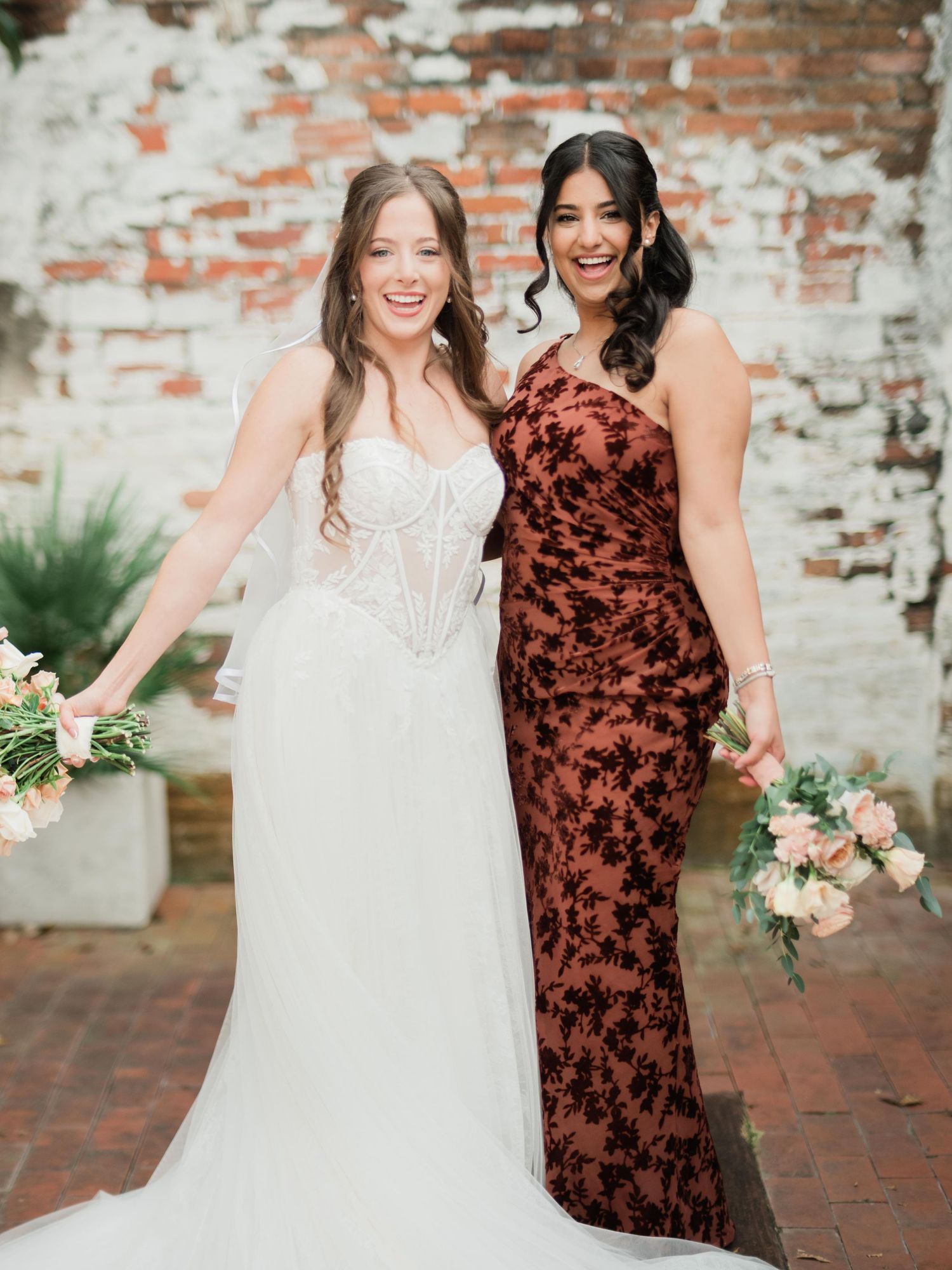Two women stand together at wedding, one wearing white bridal gown and other in burgundy velvet dress on brick patio.