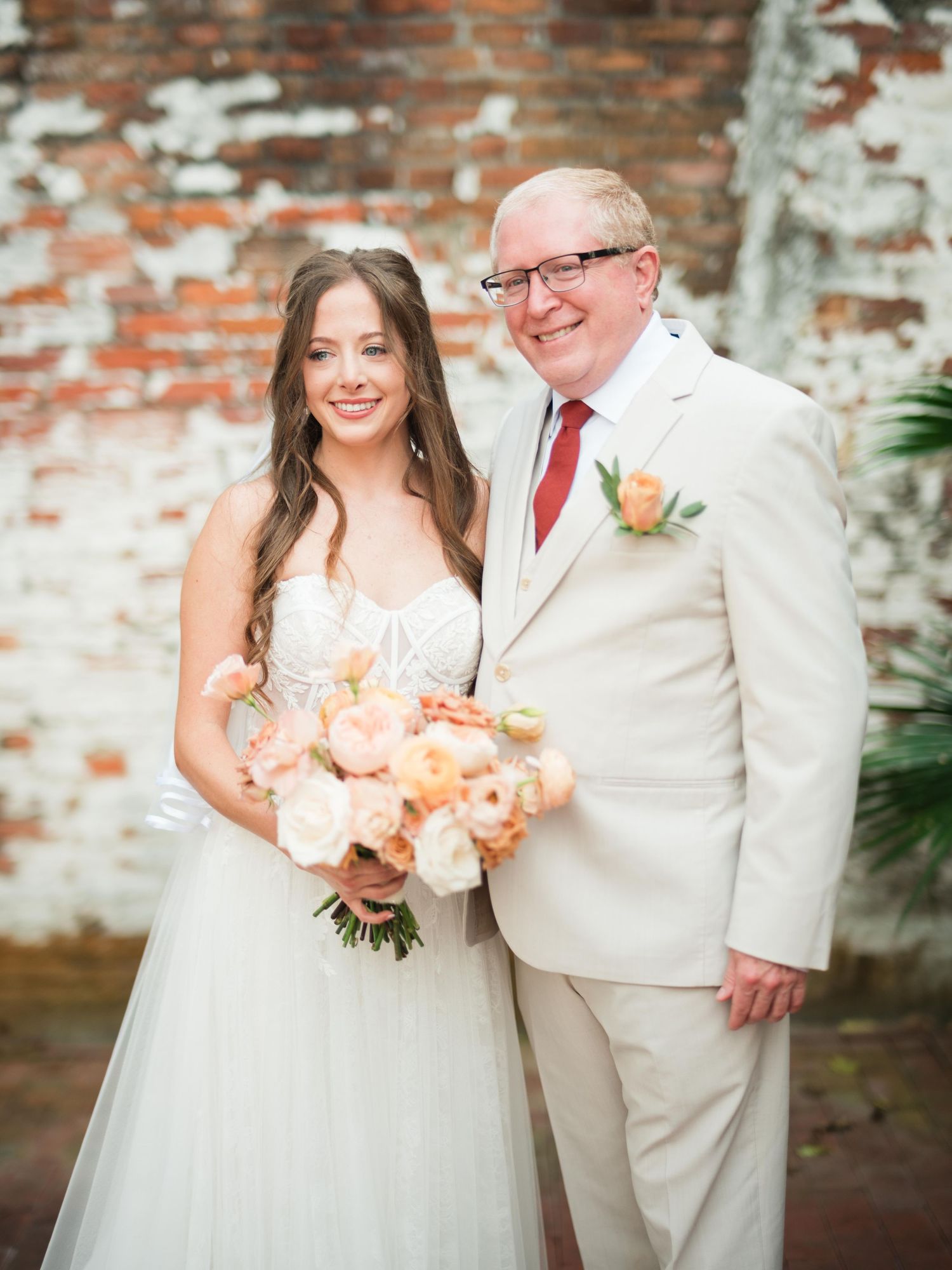 A wedding couple posing together against a rustic brick wall, with the bride holding a peach-colored floral bouquet.