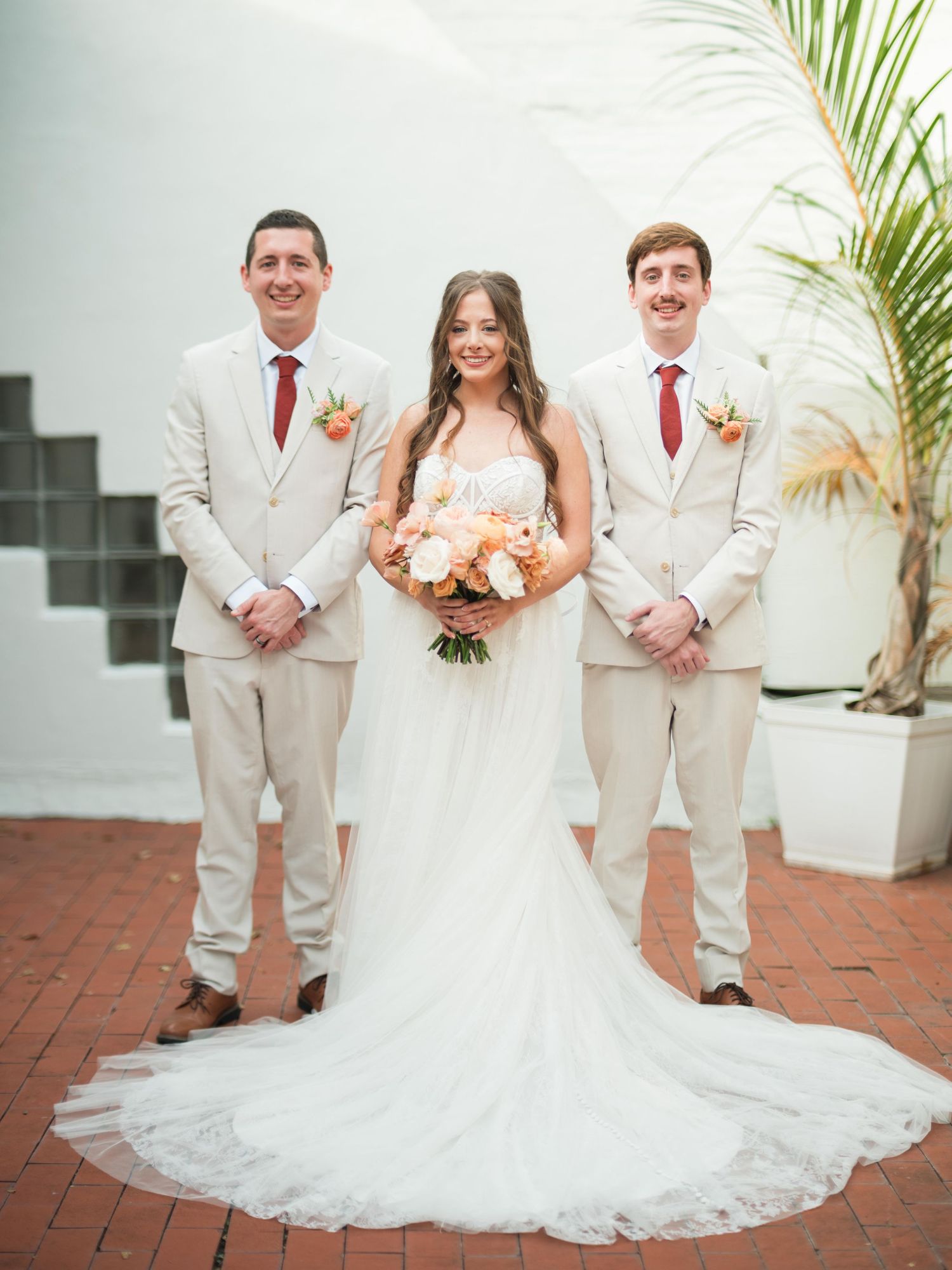 Wedding party stands on brick patio wearing cream suits coordinated with red ties and bridal gown with flowing lace train.