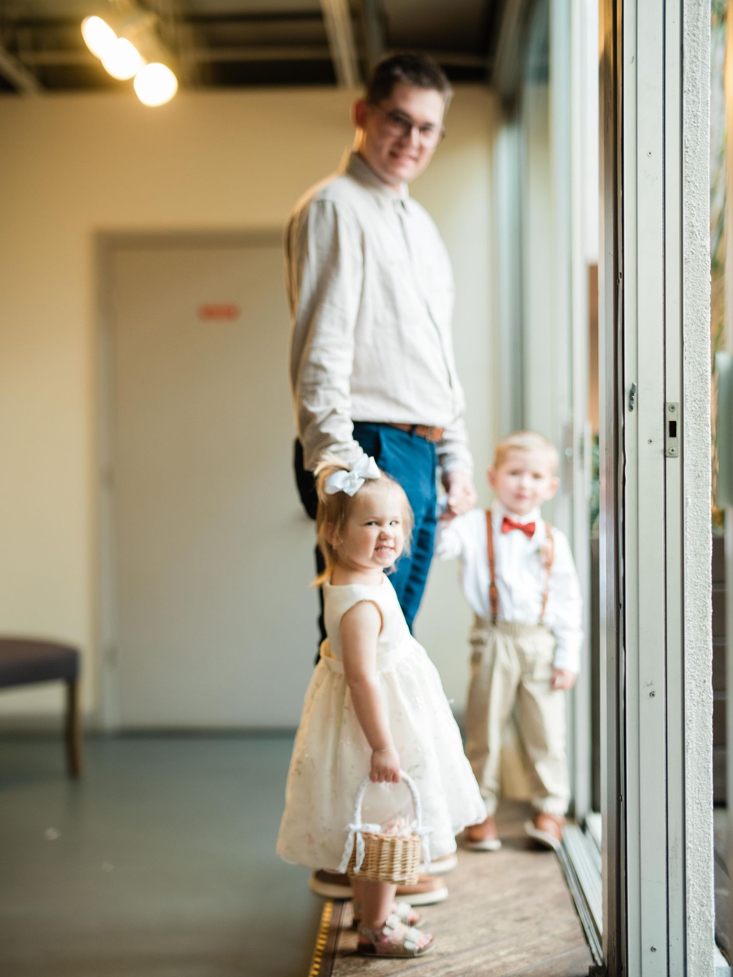 Children in formal attire stand by window in modern venue setting.