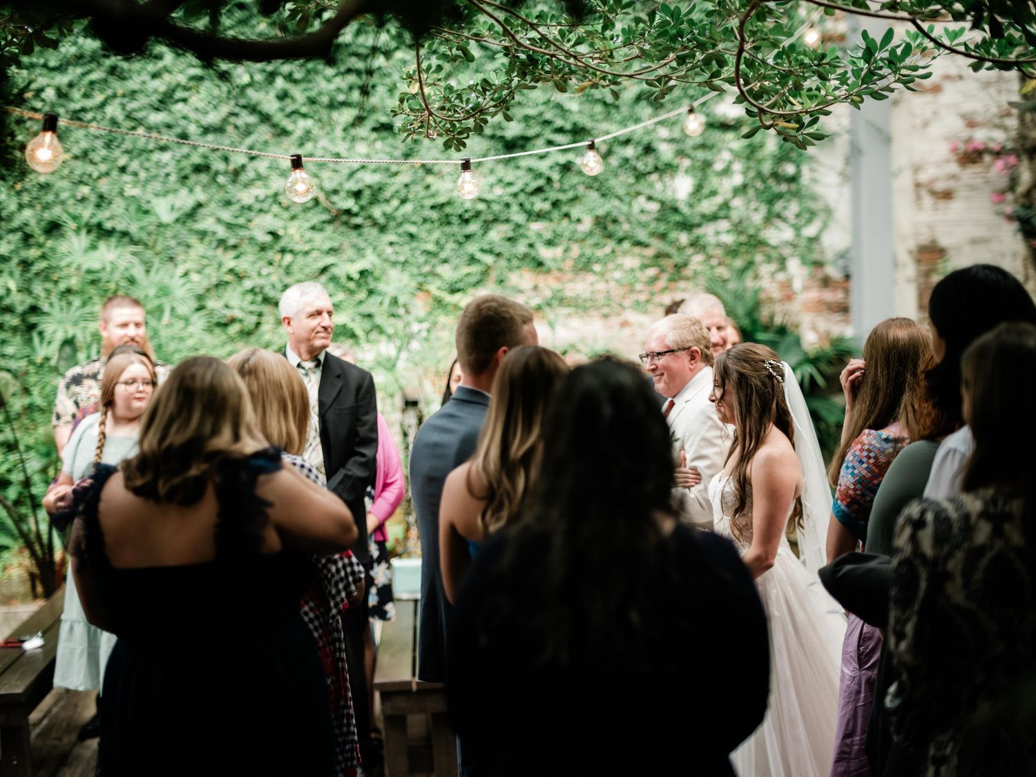 A group gathers outdoors for a ceremony surrounded by lush greenery in a backyard wedding setting.