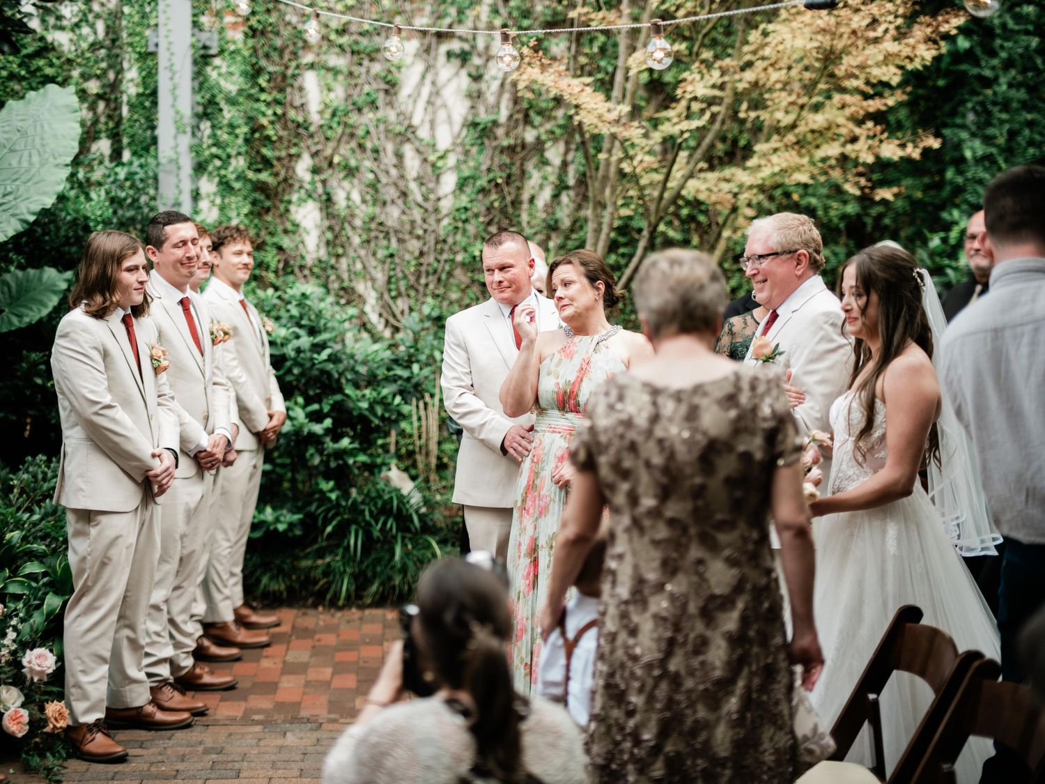 Wedding ceremony takes place in a lush garden setting with attendees wearing light beige and white attire.