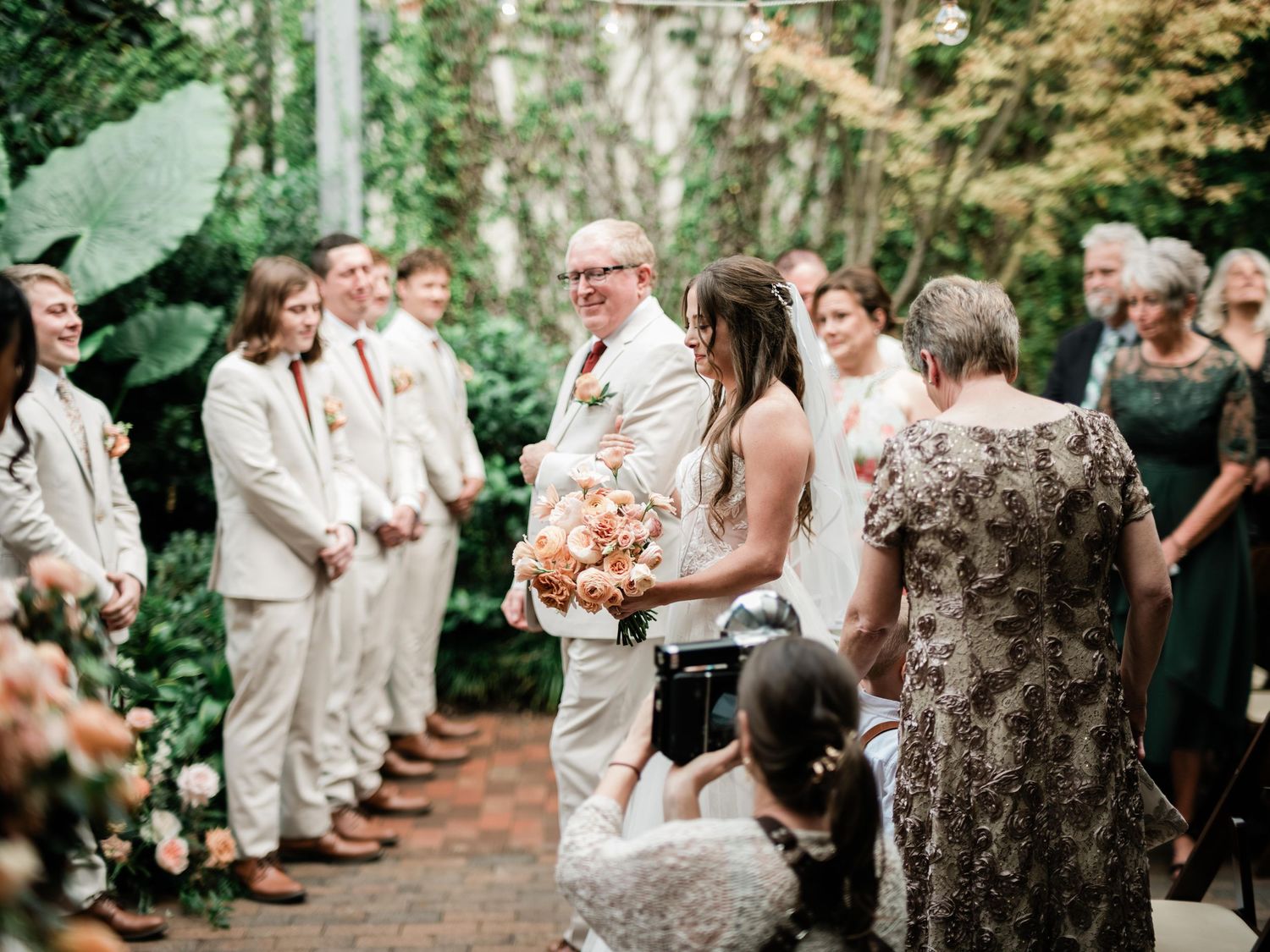 A wedding ceremony takes place in a lush garden setting with guests gathered along a brick pathway.