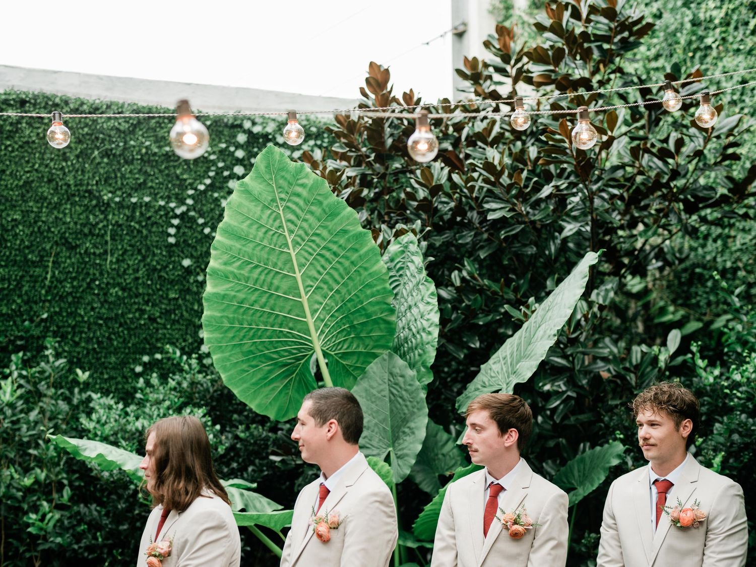 Wedding party members in light suits stand in a line against a green backdrop with string lights.