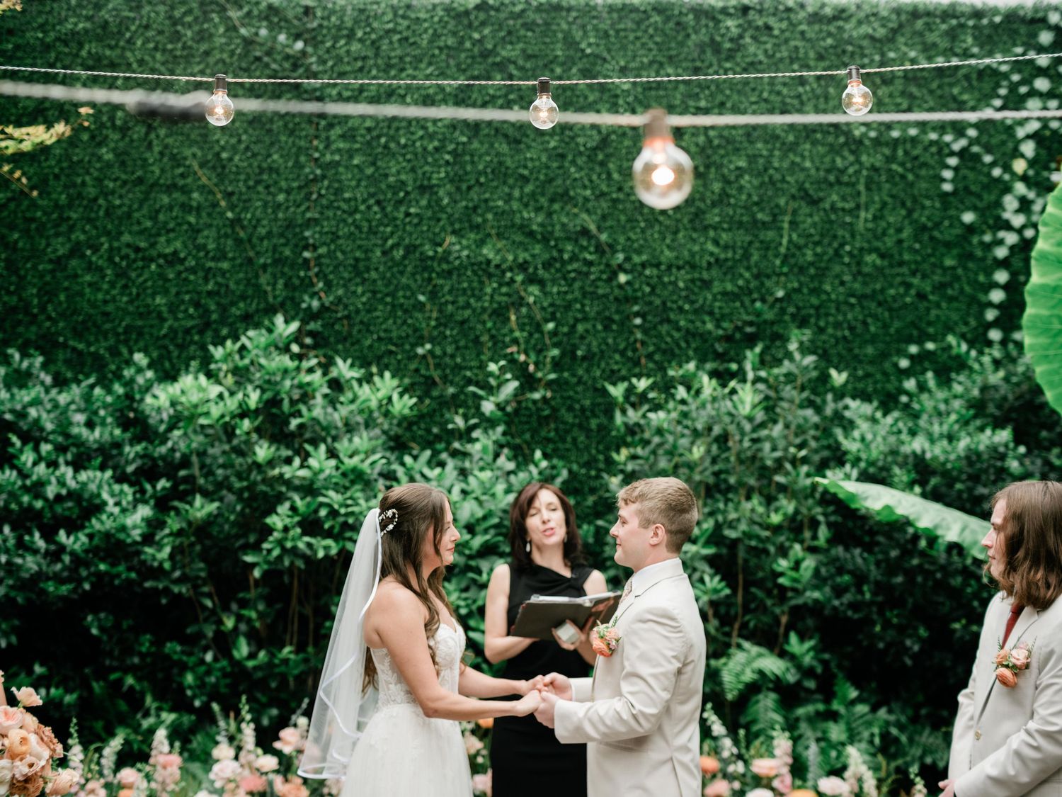 Outdoor garden wedding ceremony with string lights and greenery backdrop.