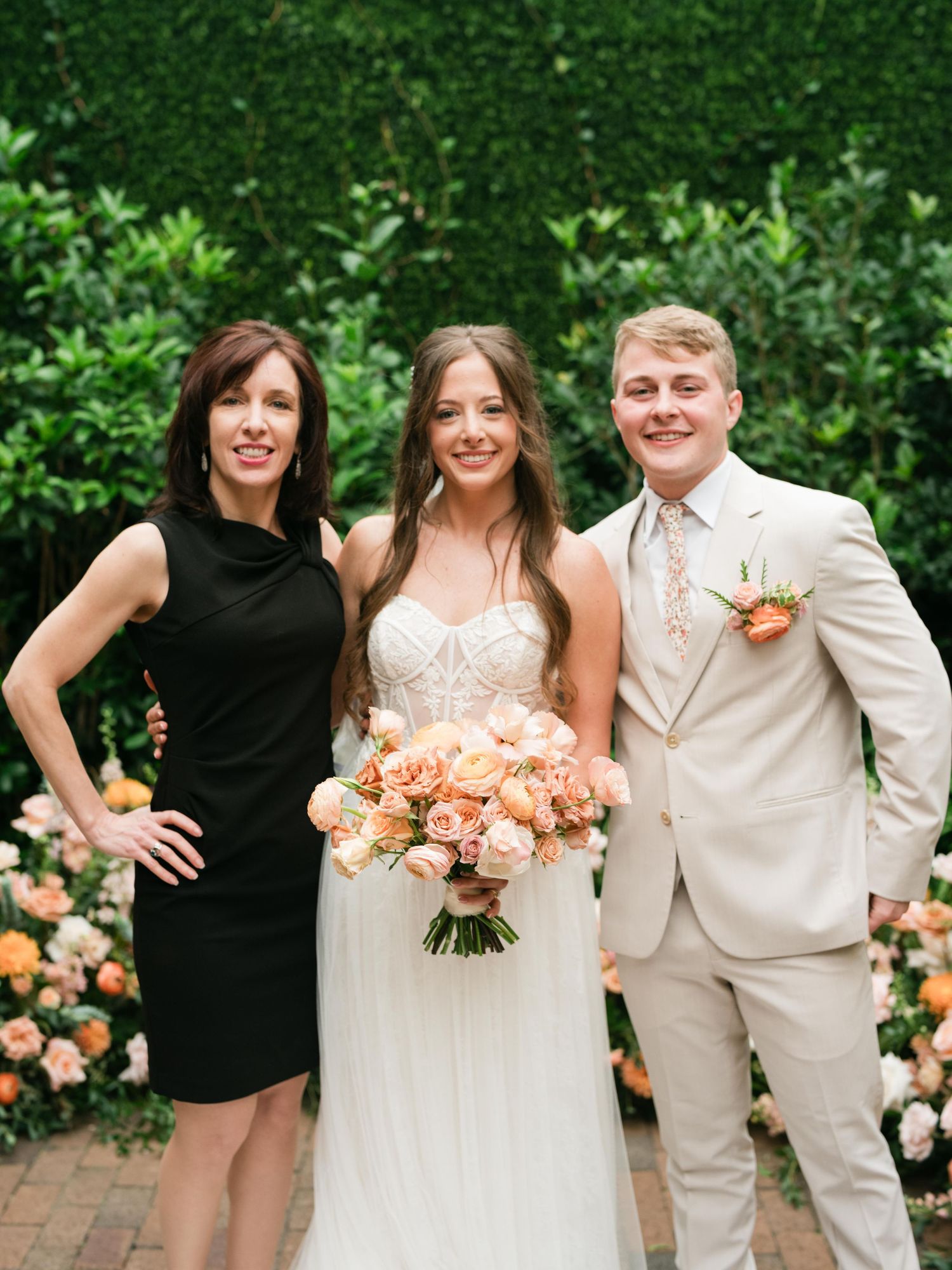 Wedding party poses against a lush green hedge backdrop with peach floral arrangements.