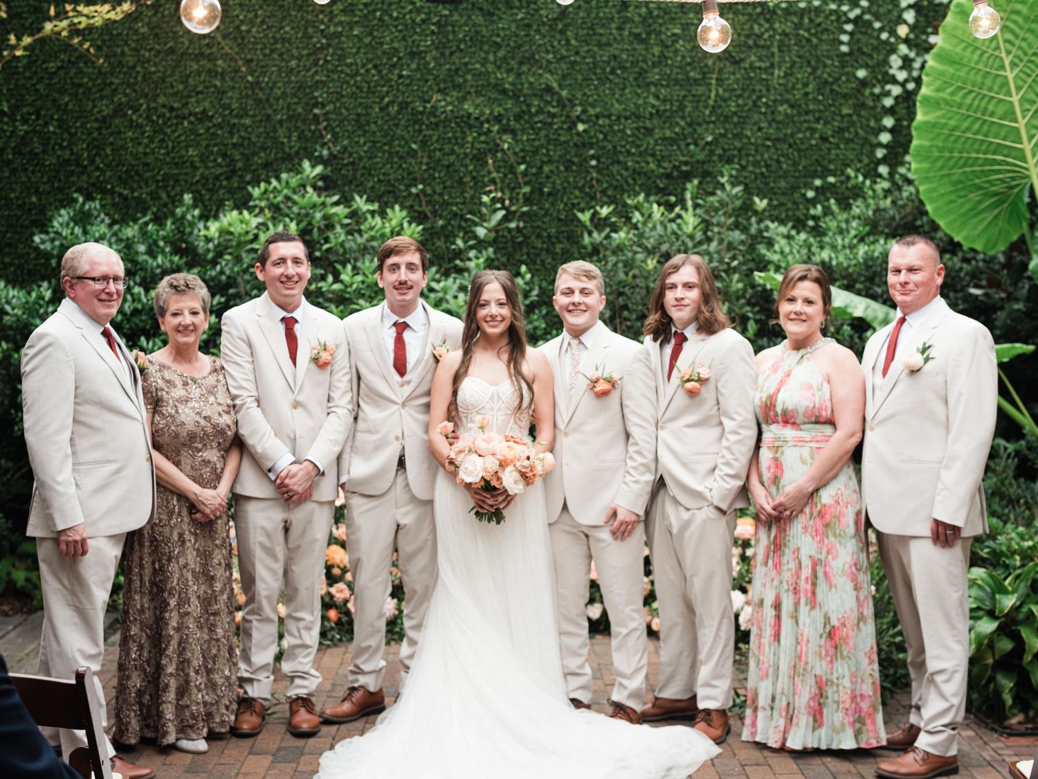Wedding party in cream suits and floral dresses pose together against a green backdrop with string lights.