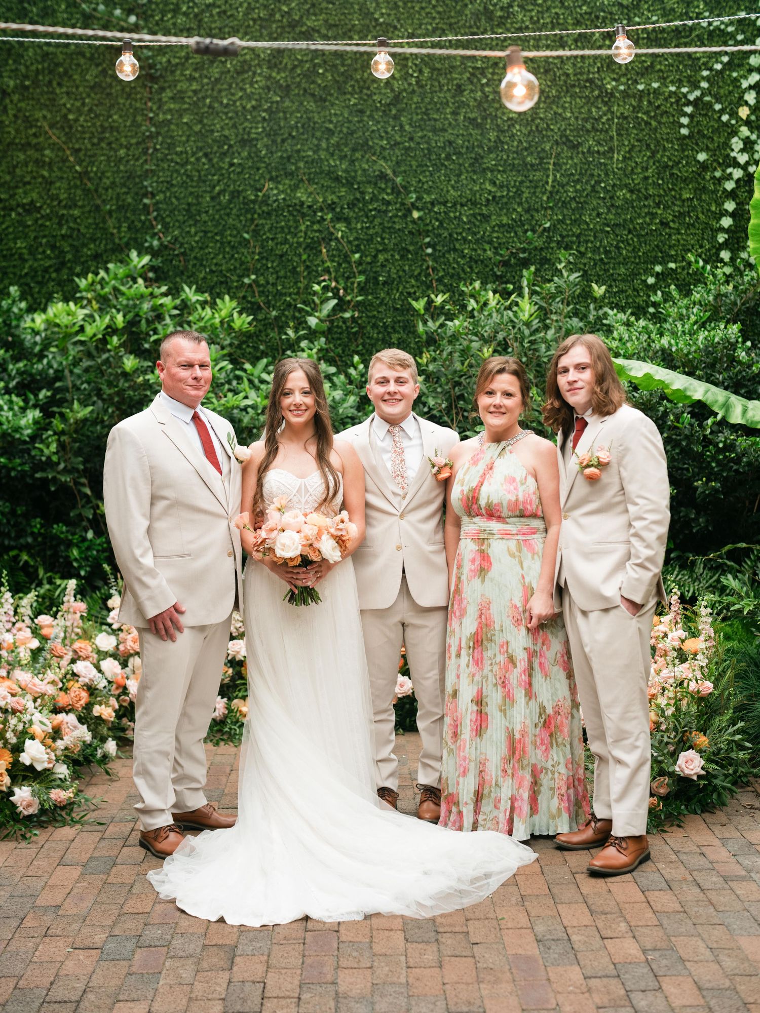 Wedding party in light beige suits and floral dresses pose against green hedge with string lights and peach flowers.