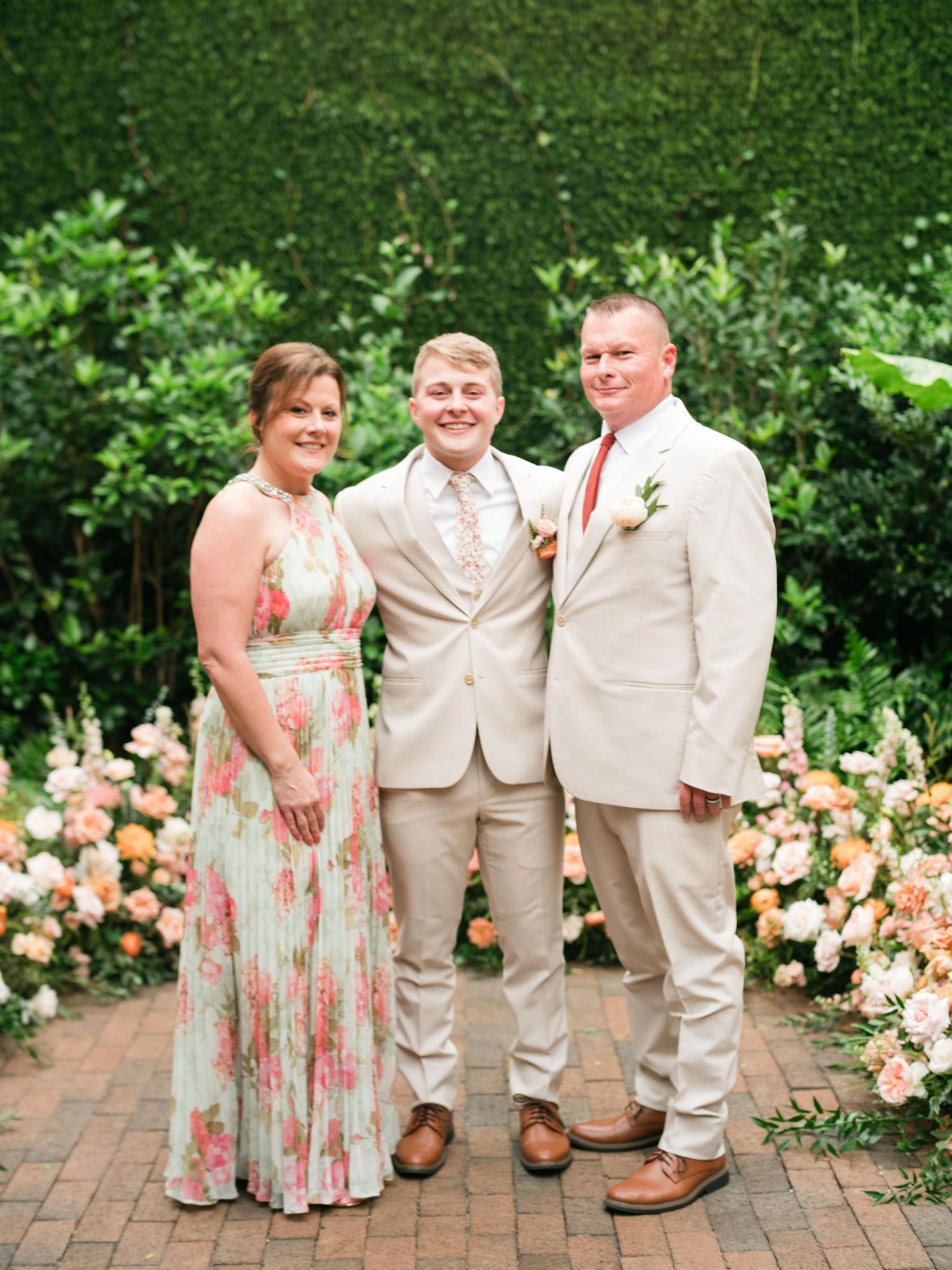 Three people in formal attire stand together on a brick path with blooming peach roses and greenery in the background.
