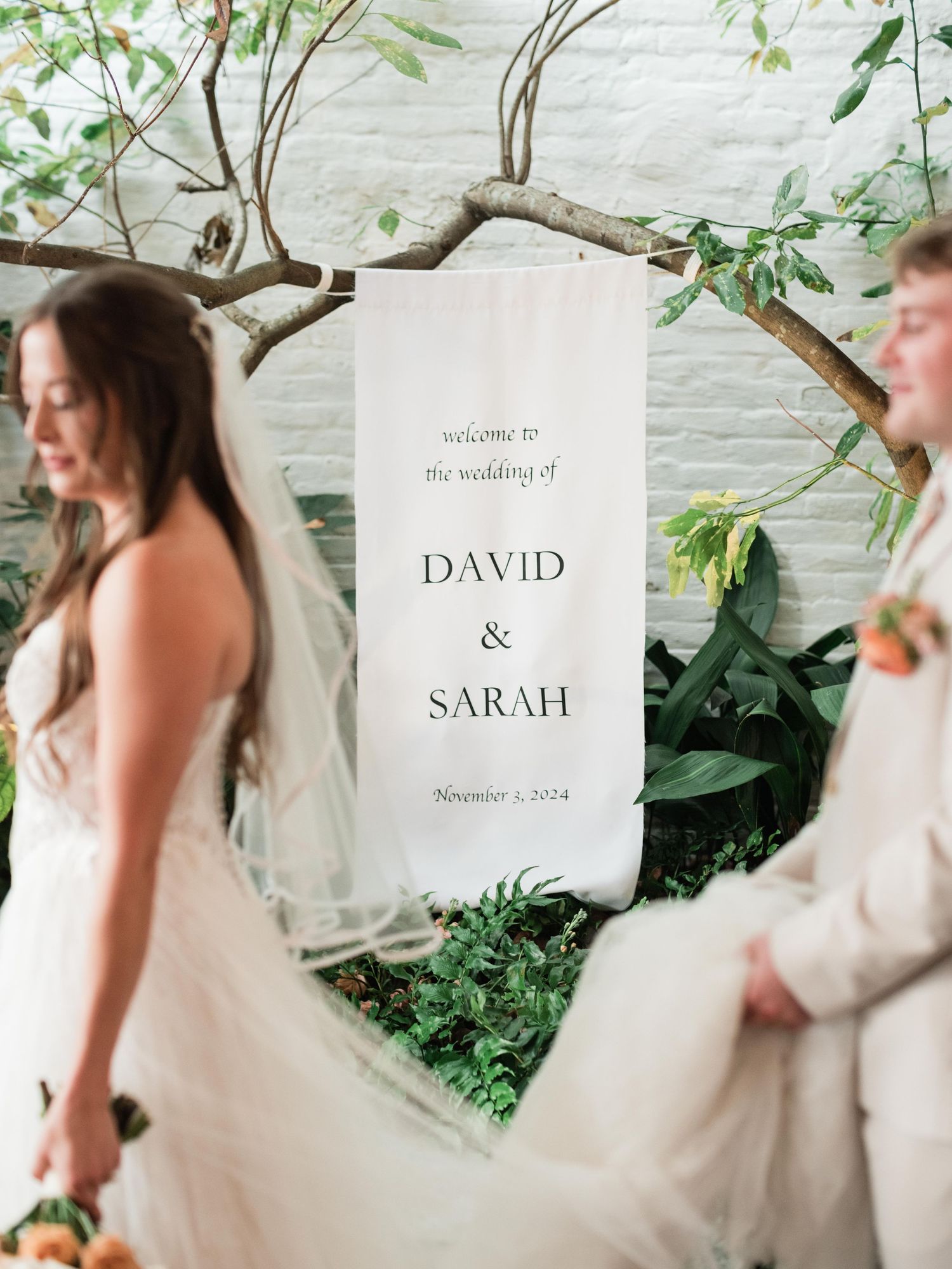 A minimalist wedding welcome sign hanging from a curved branch with greenery against a white wall.