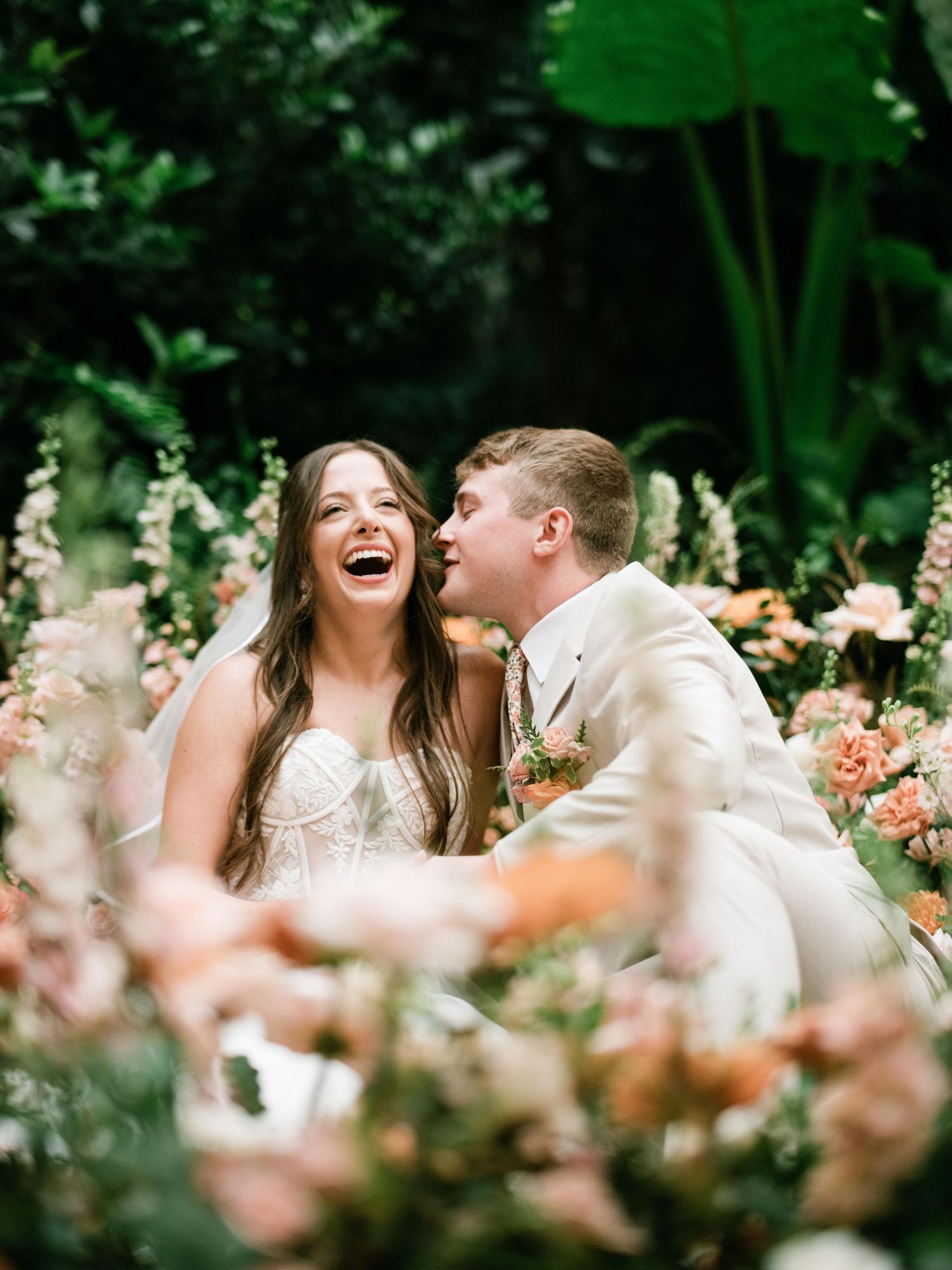Joyful moment between newlyweds surrounded by peach roses and tropical foliage.