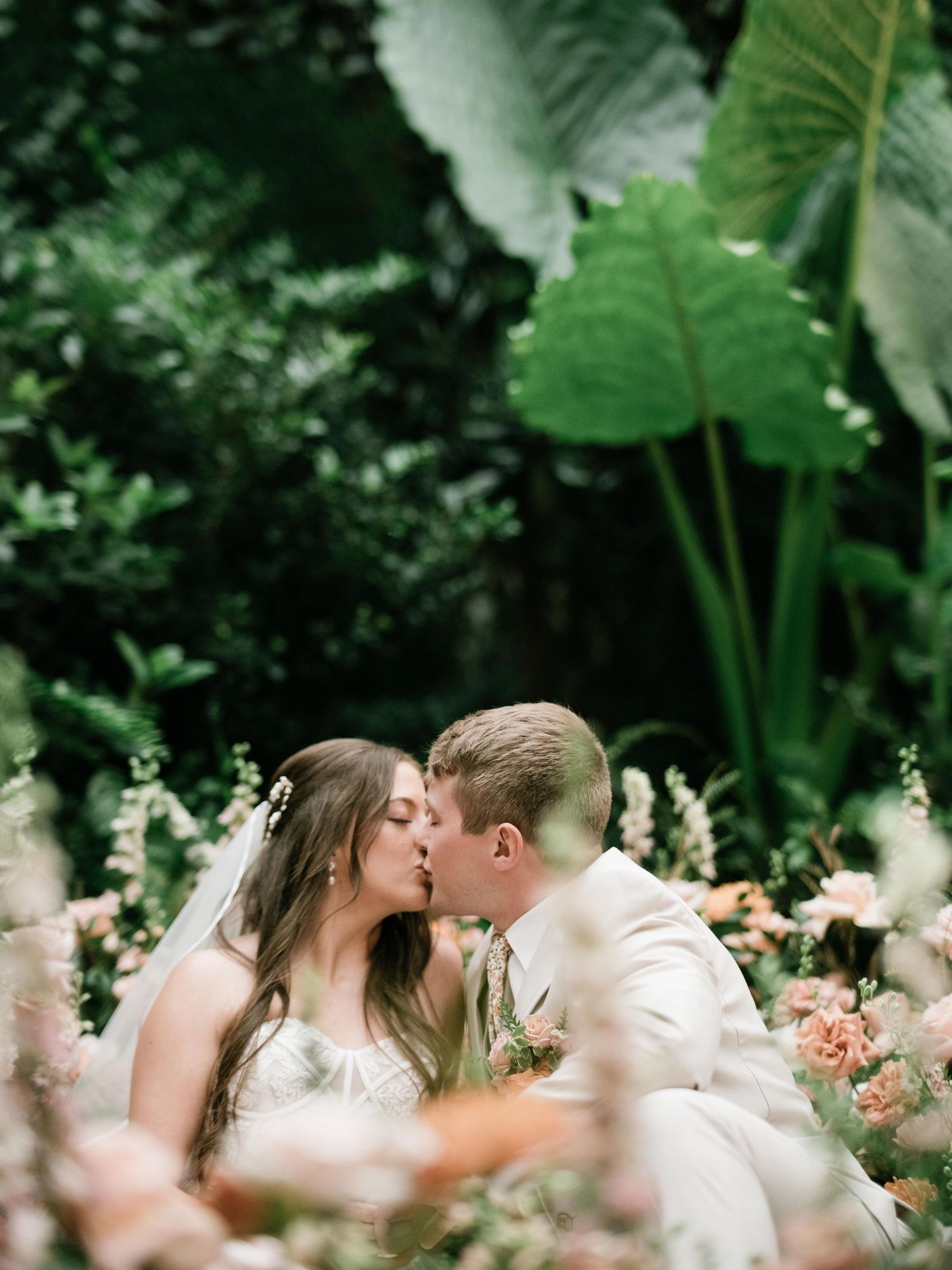 A romantic wedding couple shares a kiss among lush greenery and peachy pink flowers in a dreamy garden setting.