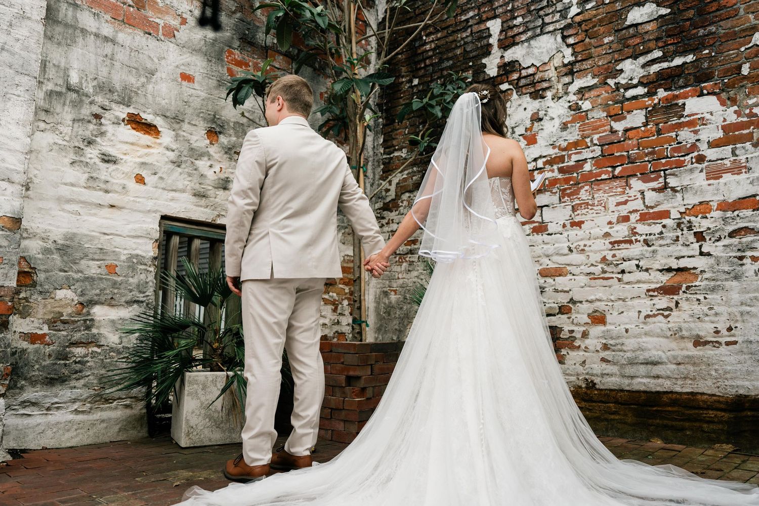 A couple in wedding attire stands against a weathered brick wall, with the bride's flowing white dress trailing behind them.