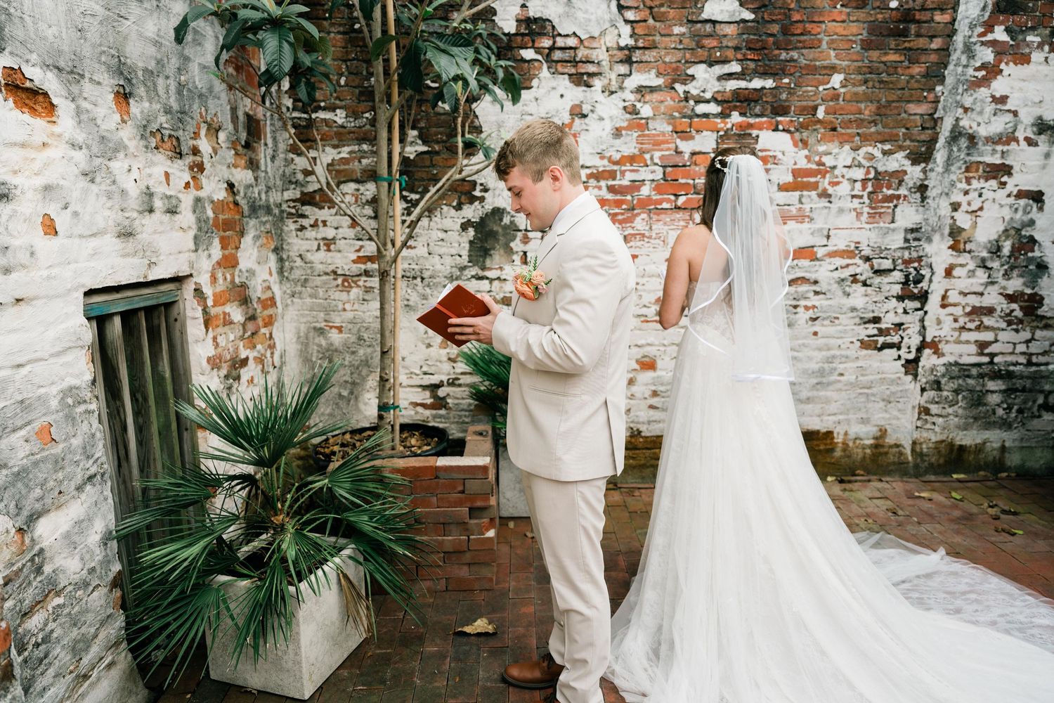 A wedding couple shares a moment near a weathered brick wall with potted palm plants.