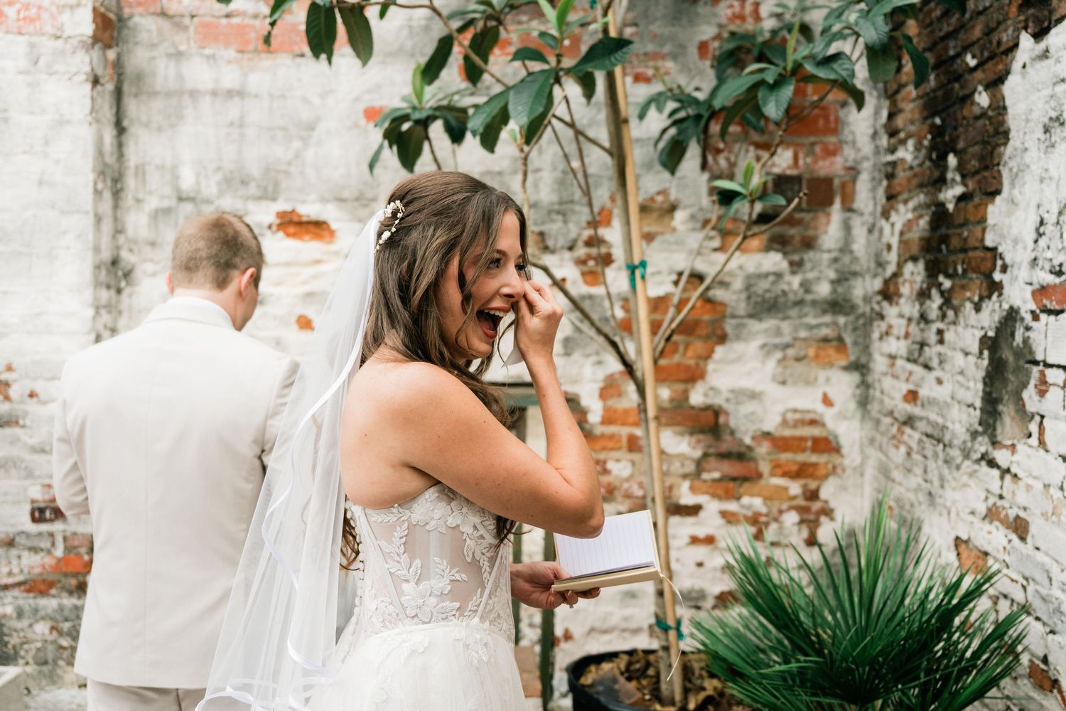 A tender wedding moment captured against rustic brick walls and greenery in a garden setting.