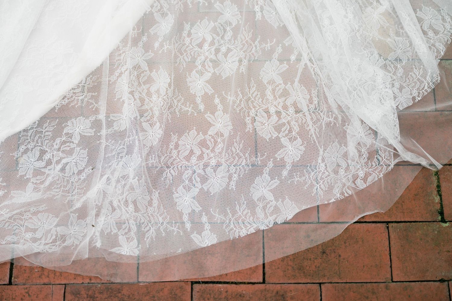 Close-up detail of delicate white lace wedding dress hem on brick pavement.