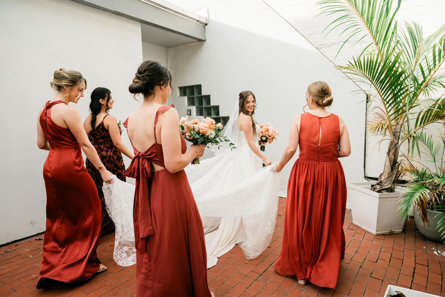 Bridesmaids in burgundy dresses help with a bride's white wedding gown on a brick patio with tropical plants.