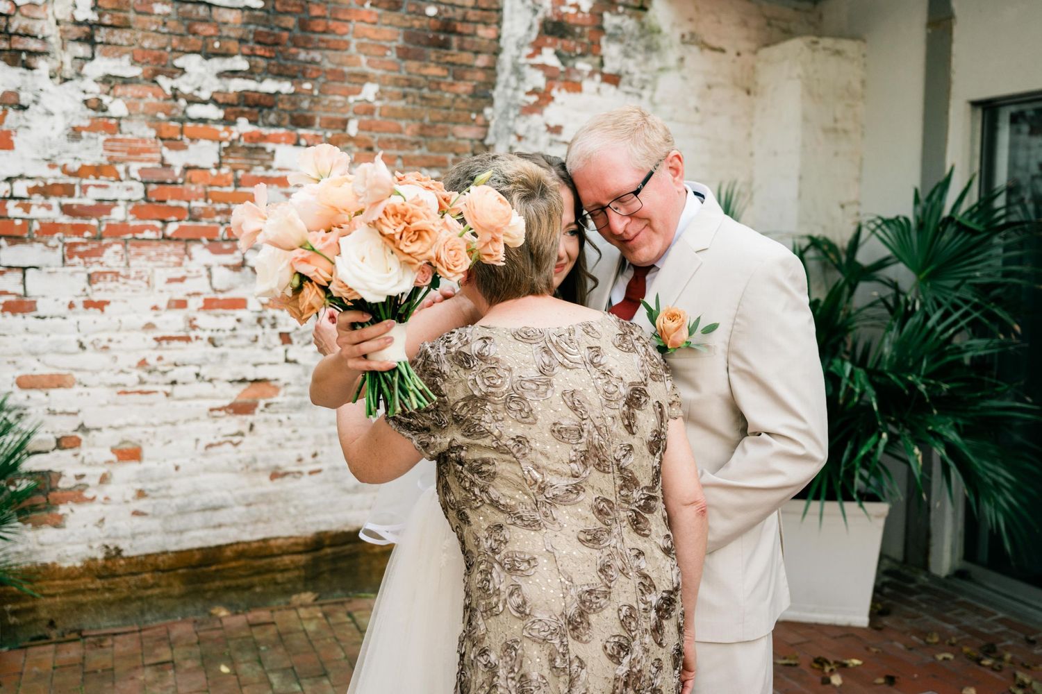 A tender moment shared between a couple in formal attire against a rustic brick wall with peach-colored flowers.