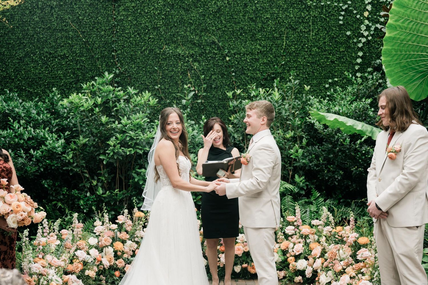 A couple exchanges vows during an outdoor wedding ceremony surrounded by lush greenery and peach florals.