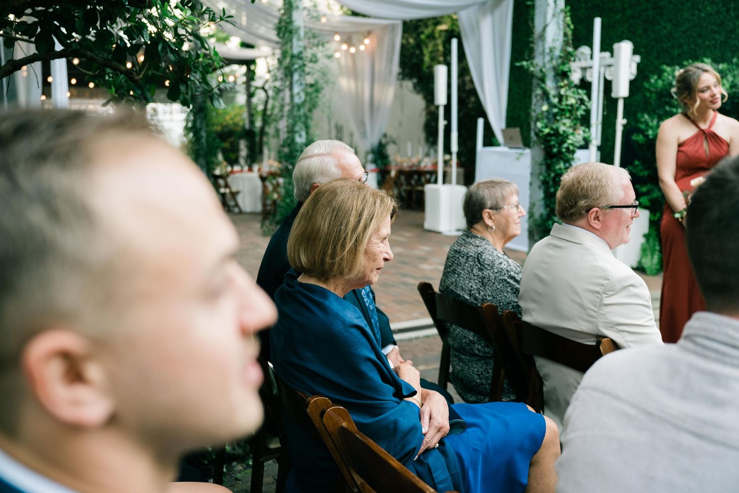 People seated in chairs at an outdoor event space with greenery and string lights in the background.