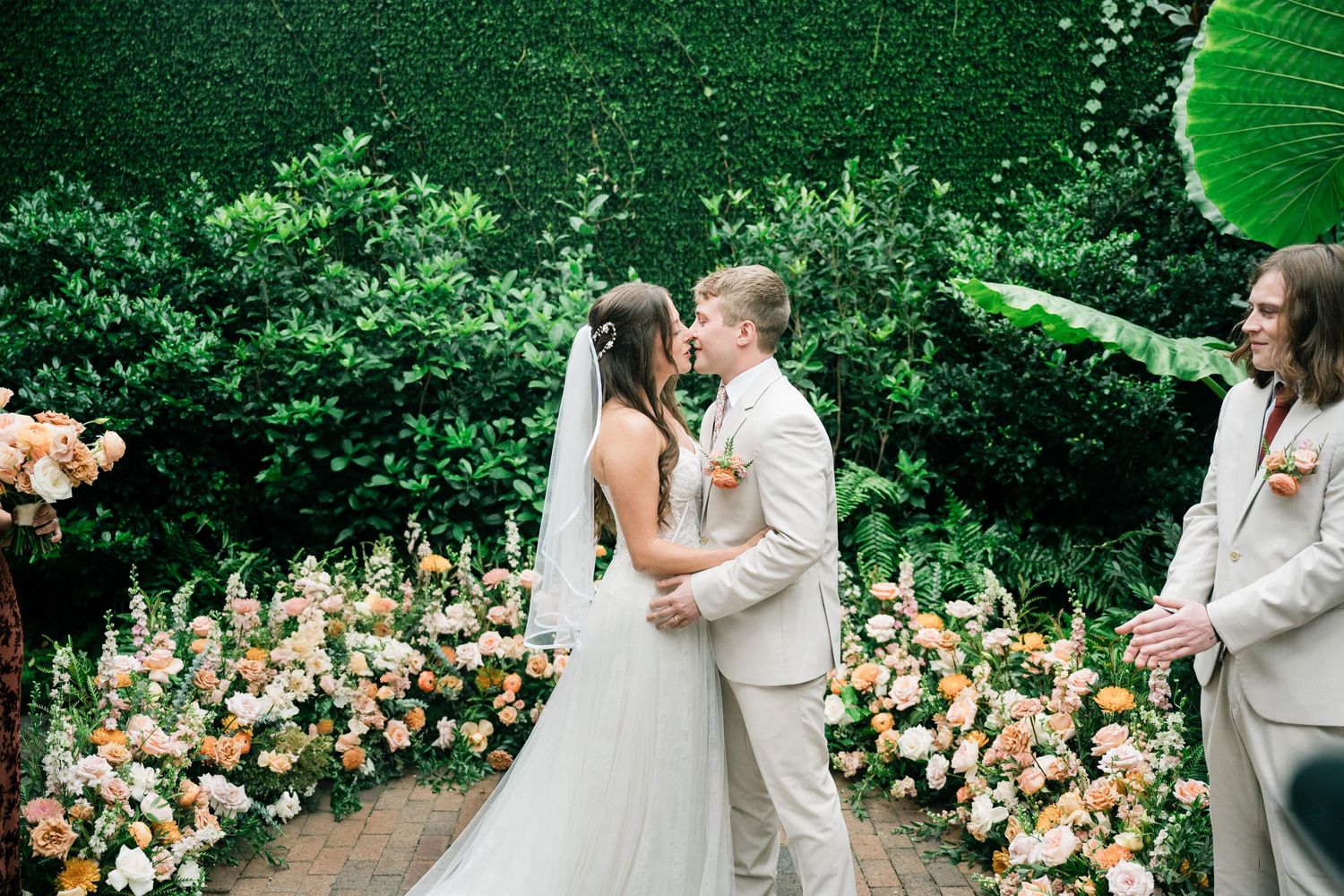 Newlyweds share their first kiss surrounded by beautiful floral arrangements in a garden setting.