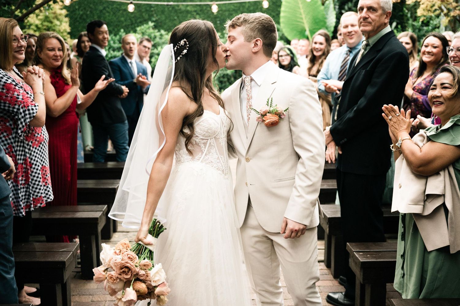 Newlyweds share their first kiss at an outdoor garden wedding ceremony as guests cheer and applaud.