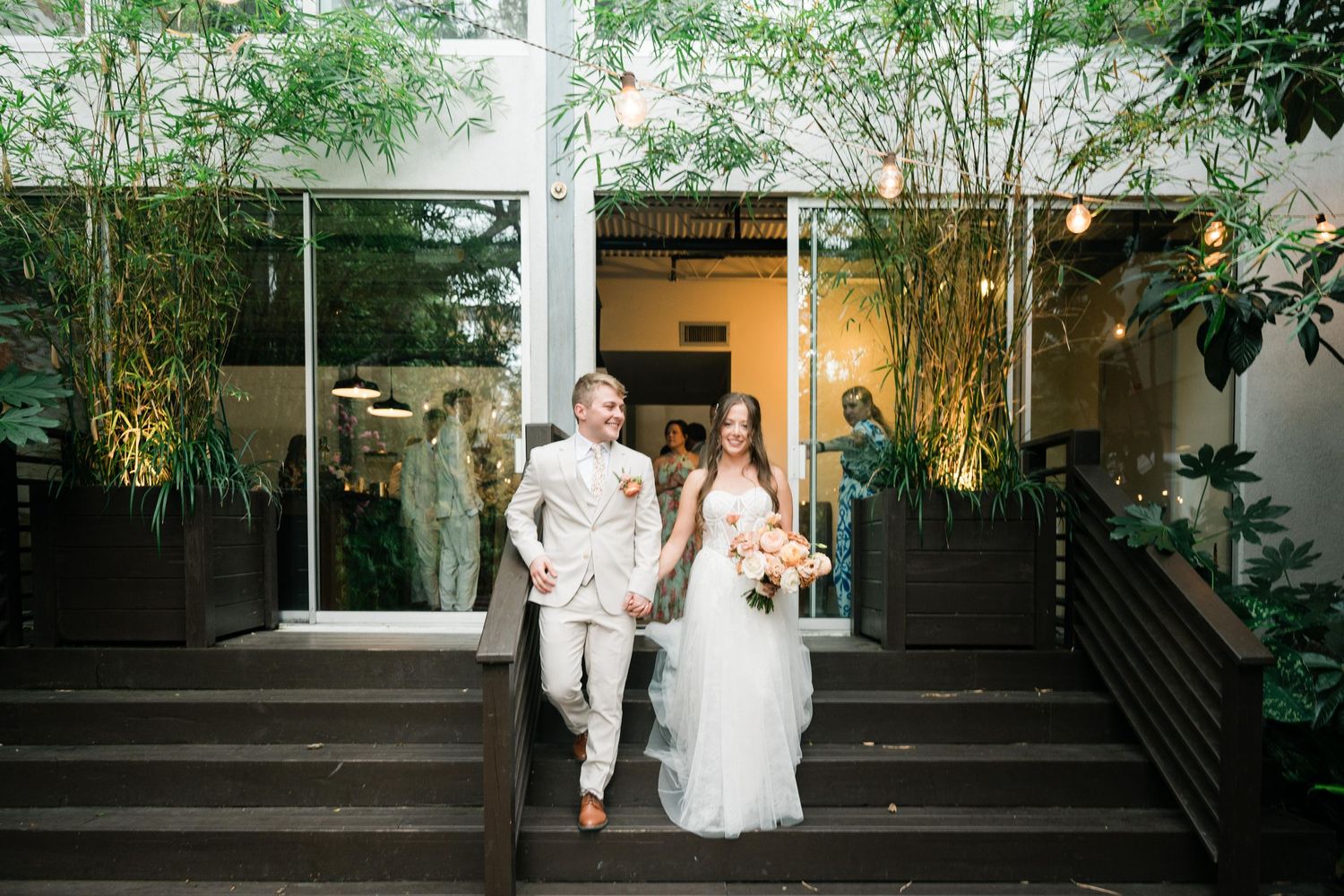 A joyful couple walks down outdoor steps surrounded by bamboo plants at a modern venue entrance.