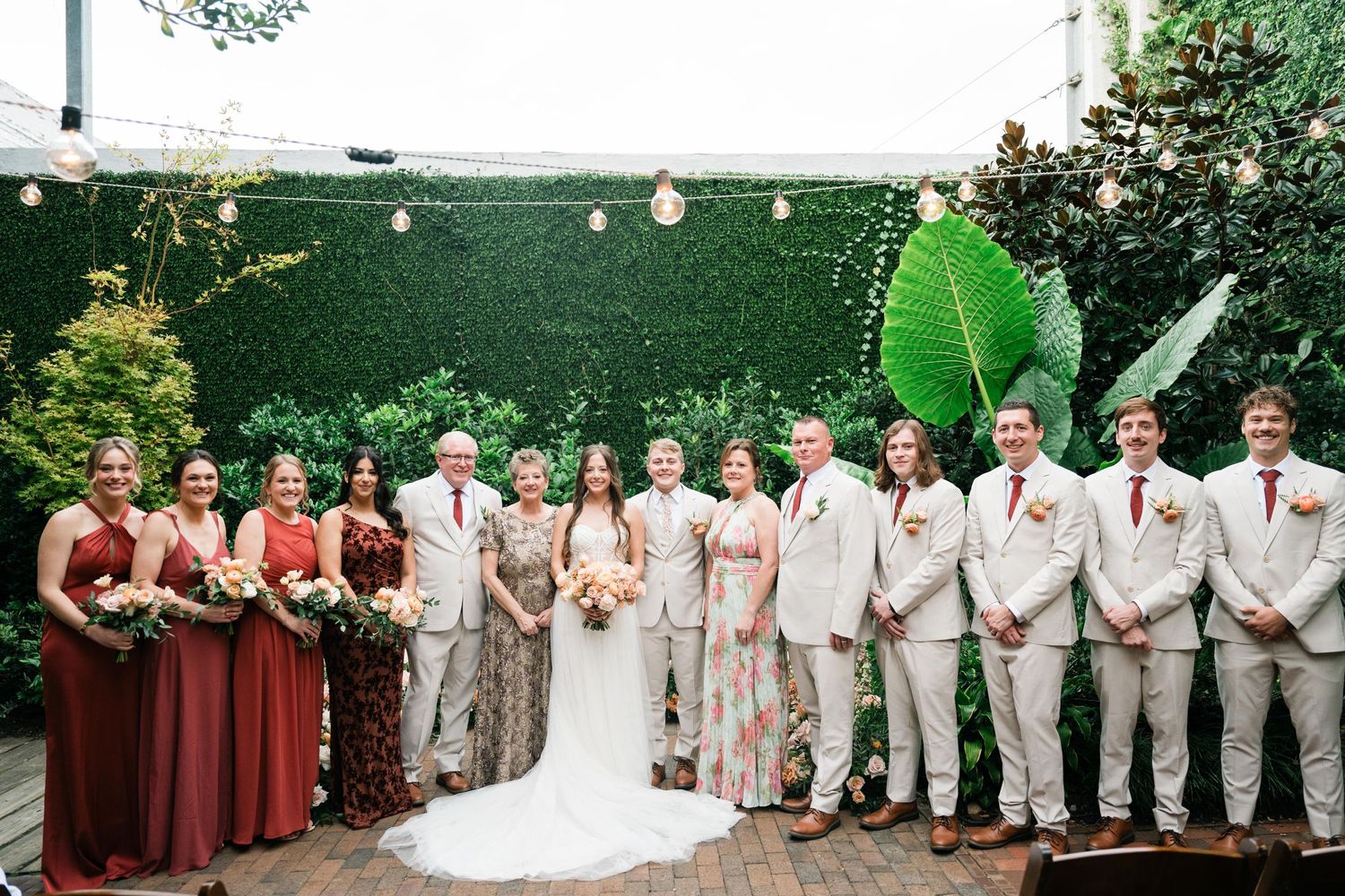 A wedding party stands in formal attire against a green wall with string lights in an outdoor venue setting.