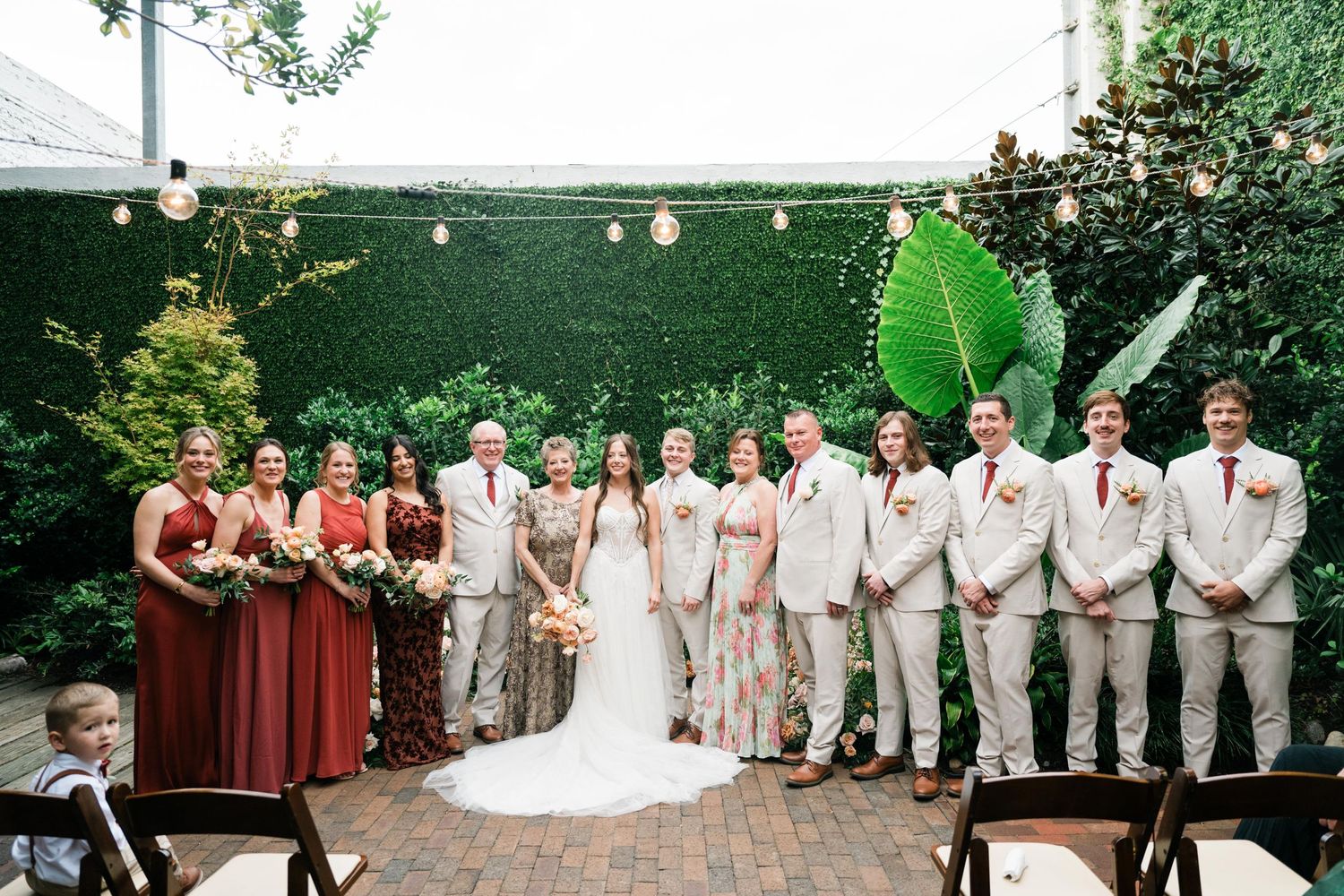 Wedding party in burgundy and neutral colors lines up against rustic outdoor garden backdrop with string lights.