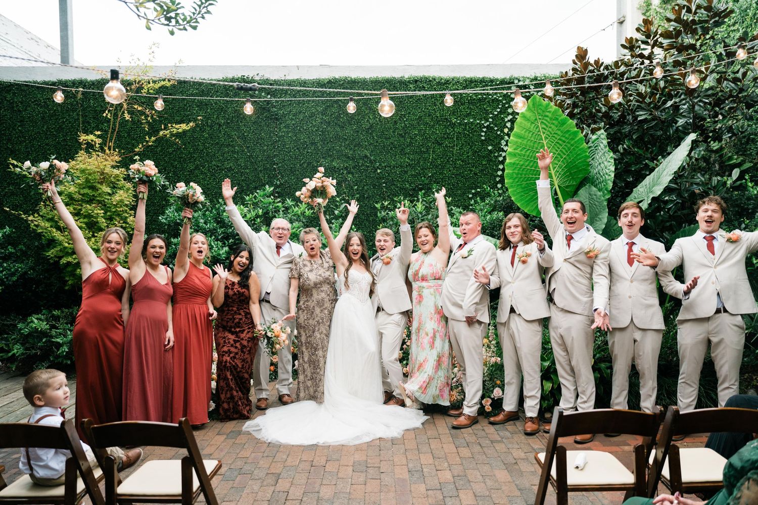Wedding party celebrates with raised arms against a hedge wall backdrop decorated with string lights and flowers.