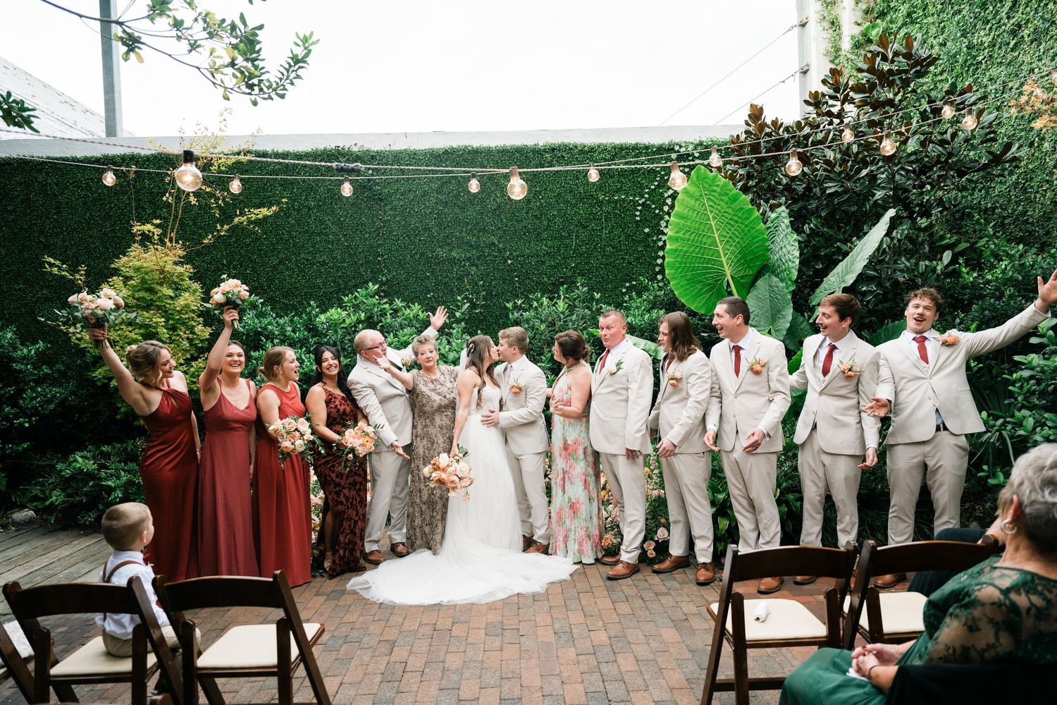 Outdoor garden wedding ceremony with bridal party in burgundy and beige standing under string lights on brick patio.