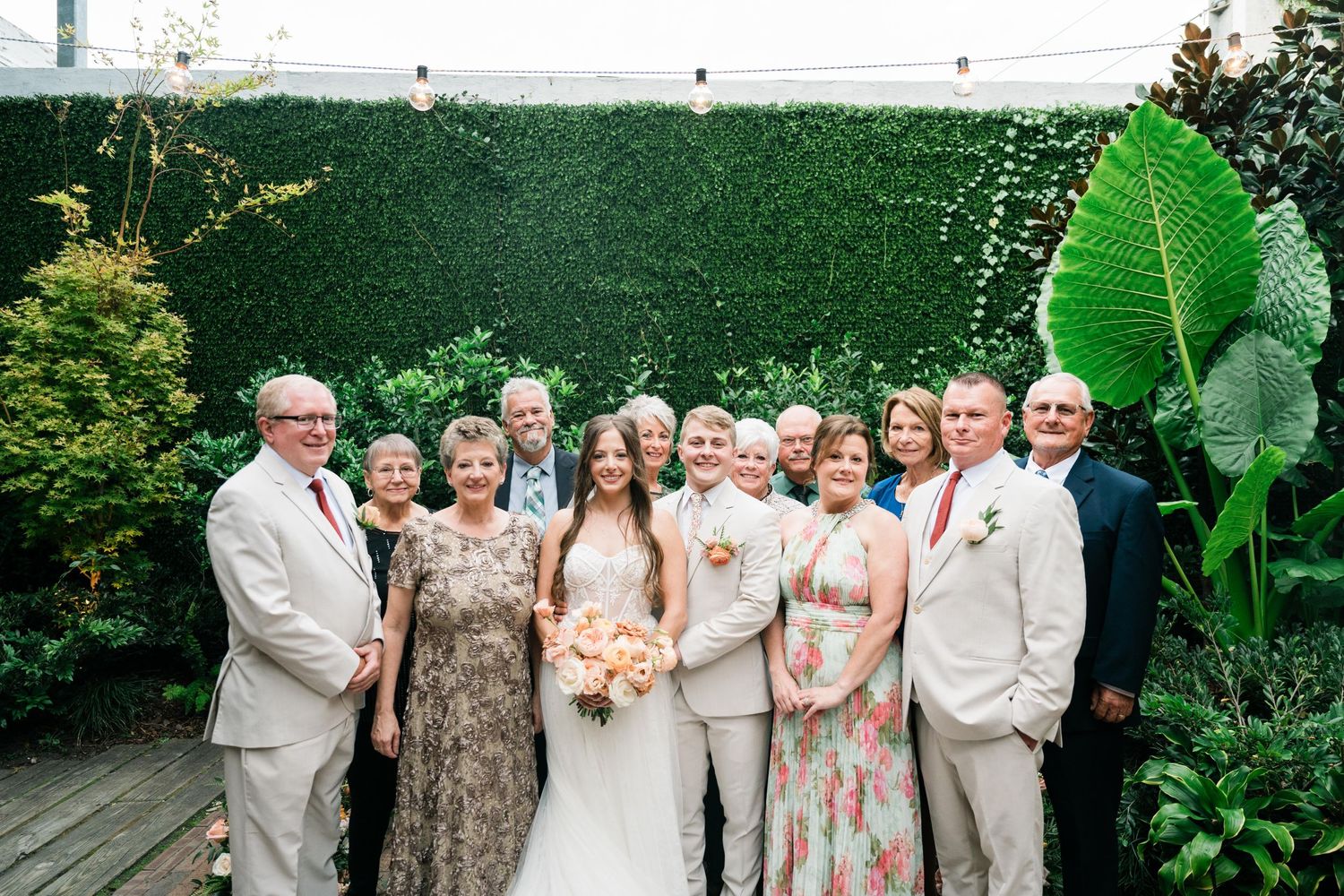 Wedding party in light colored attire poses together against a lush green hedge wall with tropical plants.