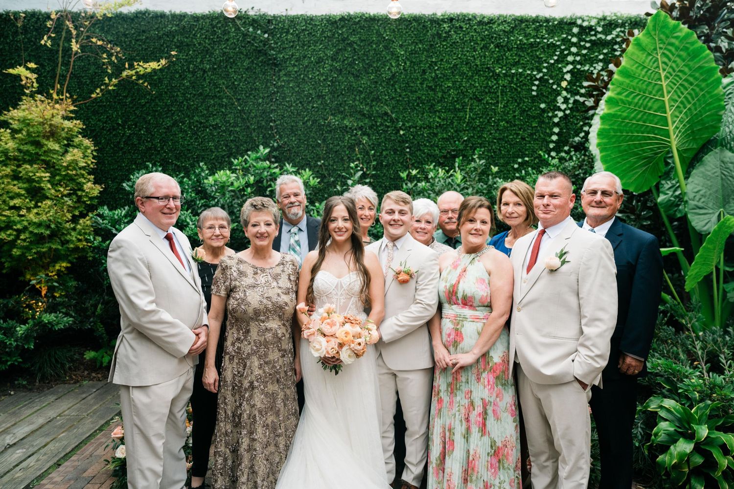 A wedding party in light summer attire poses together in front of a lush green hedge backdrop in a garden setting.