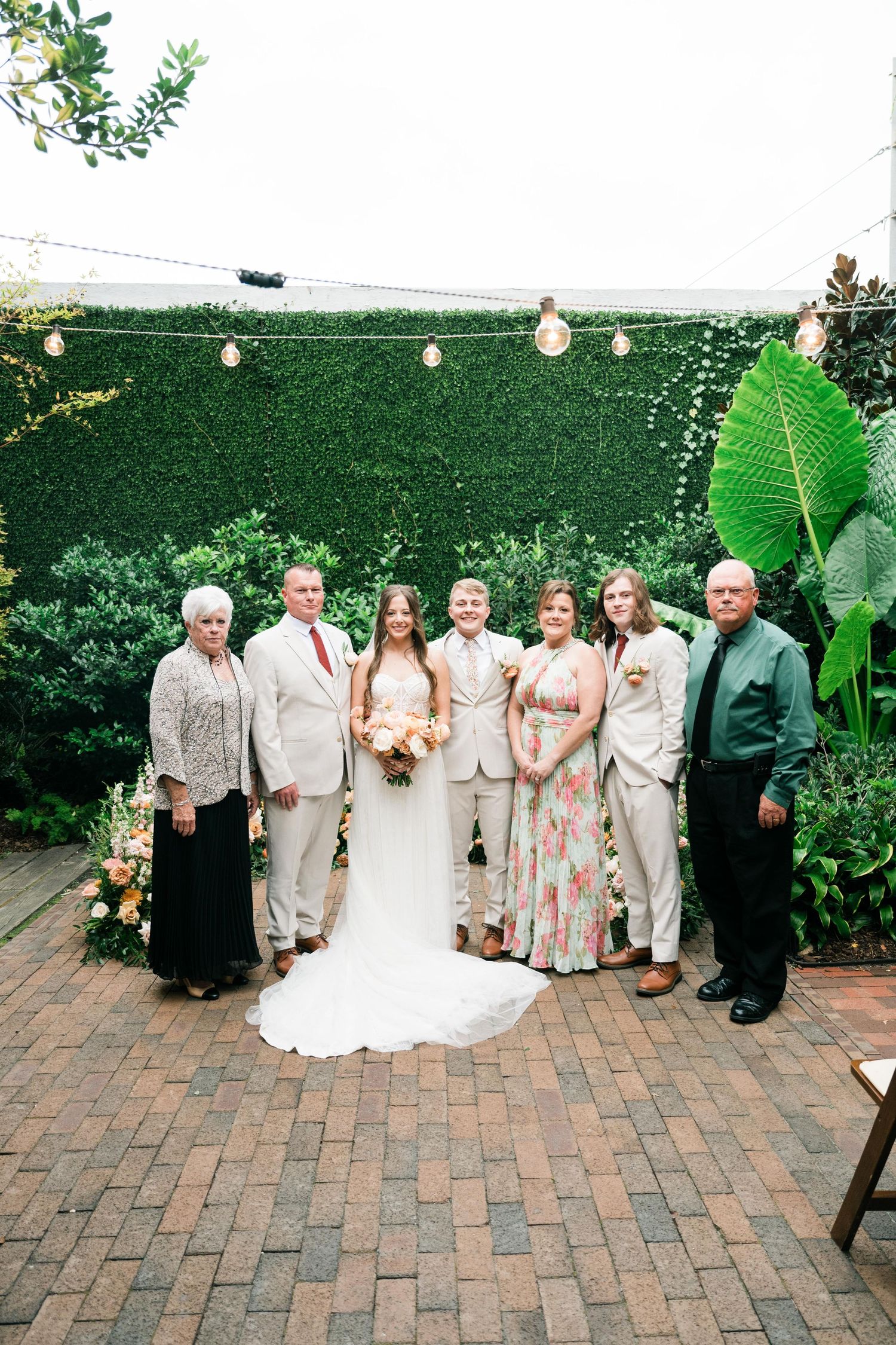 Wedding group photo in lush garden courtyard with string lights overhead and brick patio, featuring pastel and neutral attire.