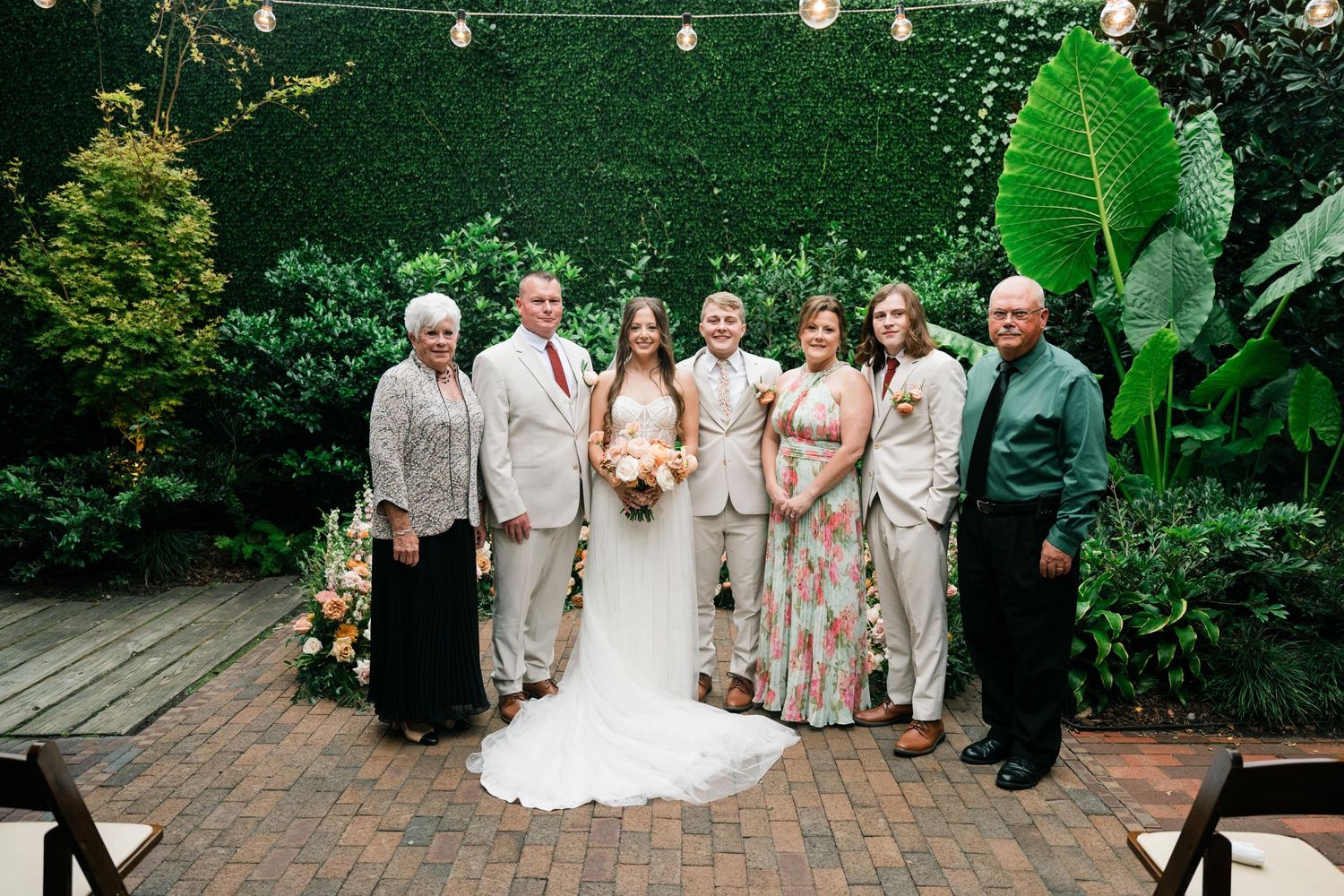 Wedding party poses on a brick patio with lush greenery and hanging lights in the background.